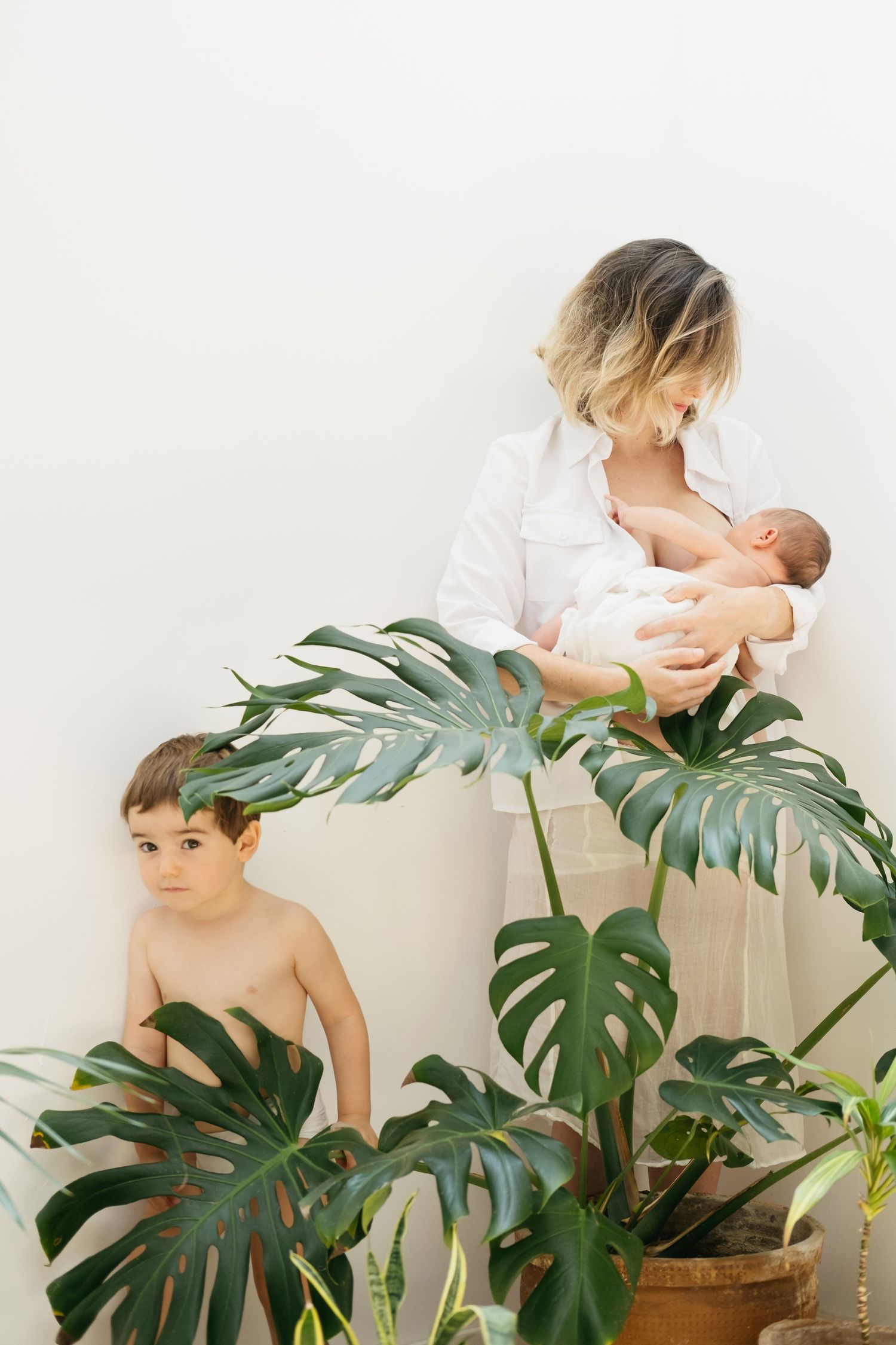 Imagen familiar de una madre dando de mamar a su bebé recién nacido junto a su hijo mayor que se esconde tras una planta monstera deliciosa