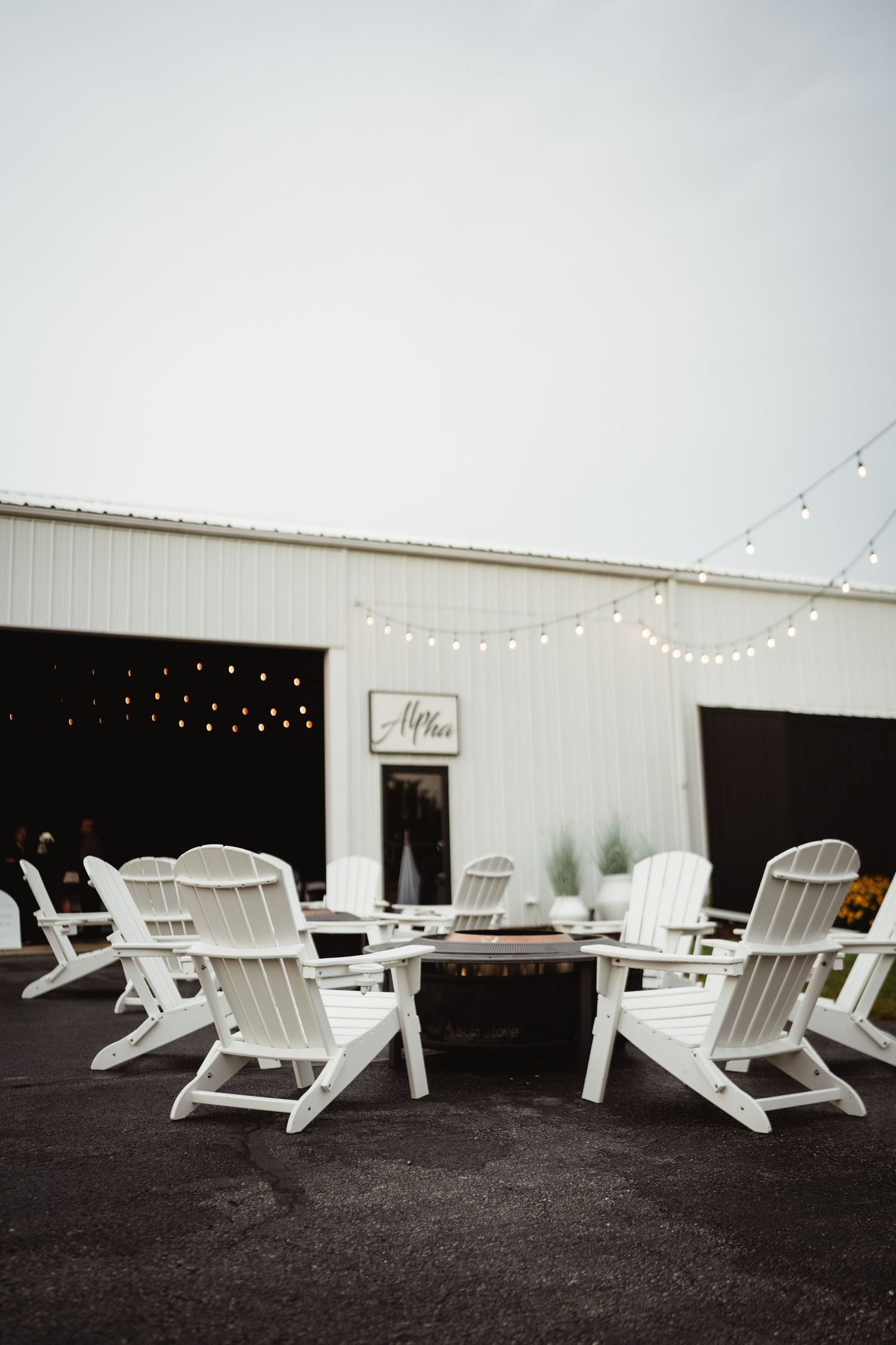 White Adirondack chairs arranged around a modern patio space with string lights and black accents at outdoor venue.