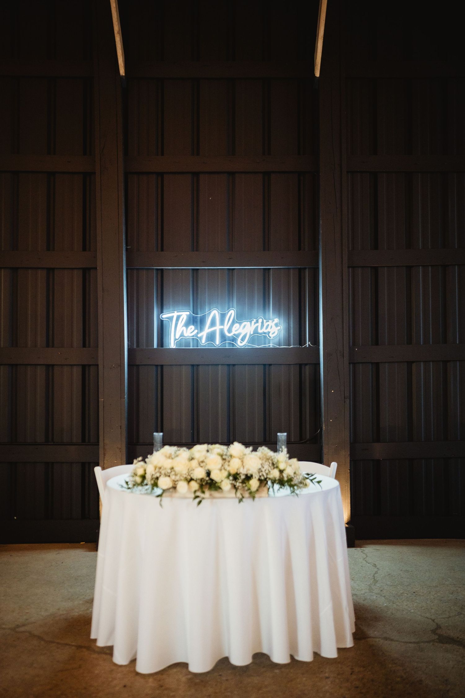 Round reception table with white tablecloth and floral centerpiece illuminated by neon sign in dark wooden venue.