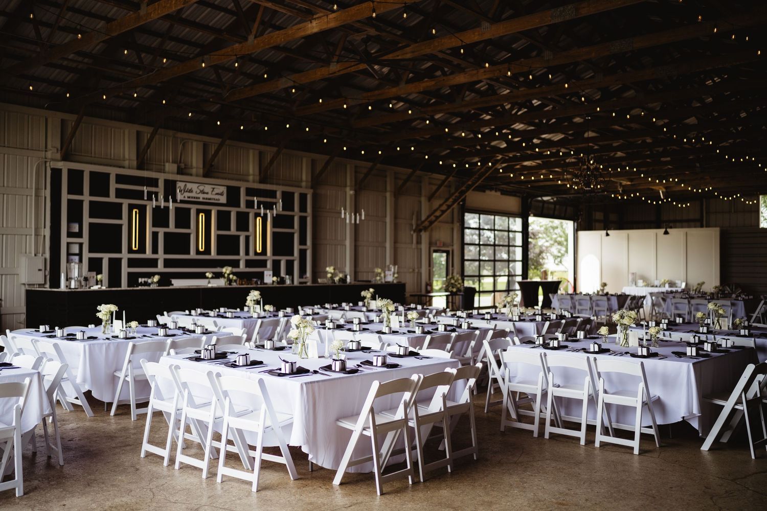 Large indoor wedding reception venue setup with white folding chairs and tables set in rows under rustic wooden beams.