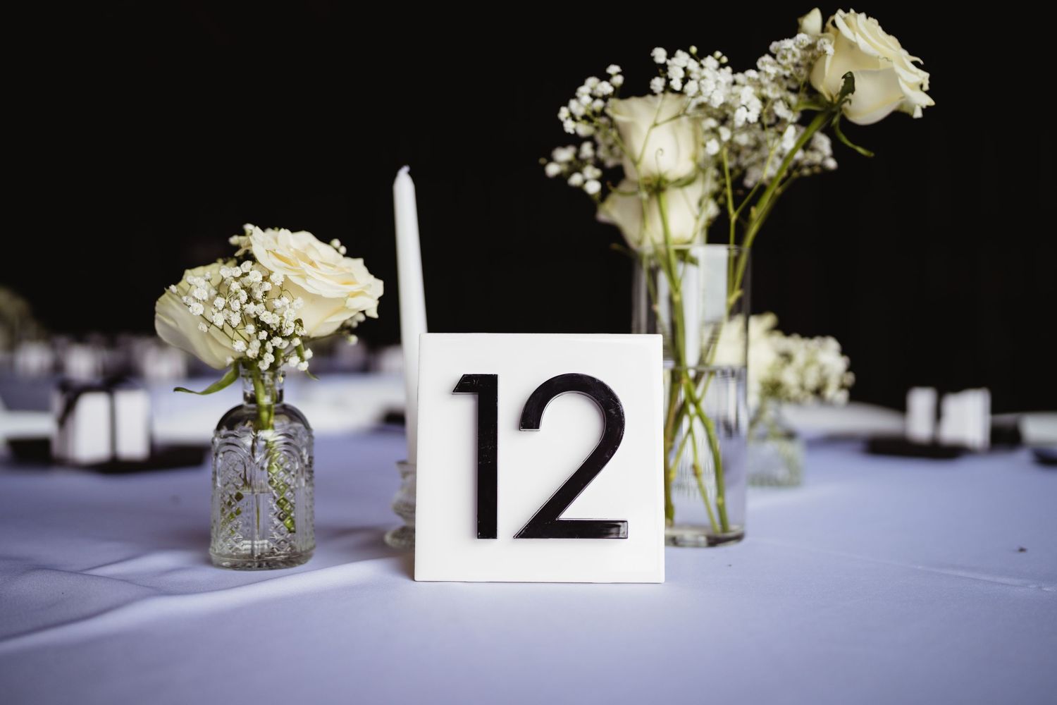 A table number 12 display with white roses and baby's breath in glass vases on a blue tablecloth against dark background.