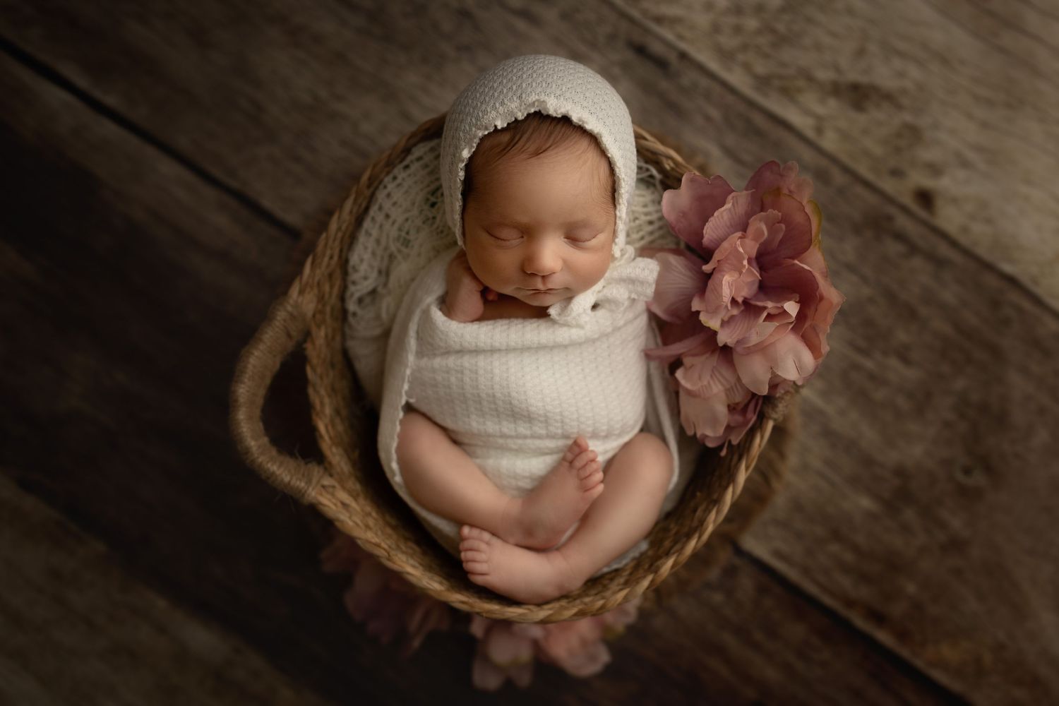 Newborn photography featuring baby in white wrap with pink flower in rustic basket setting.