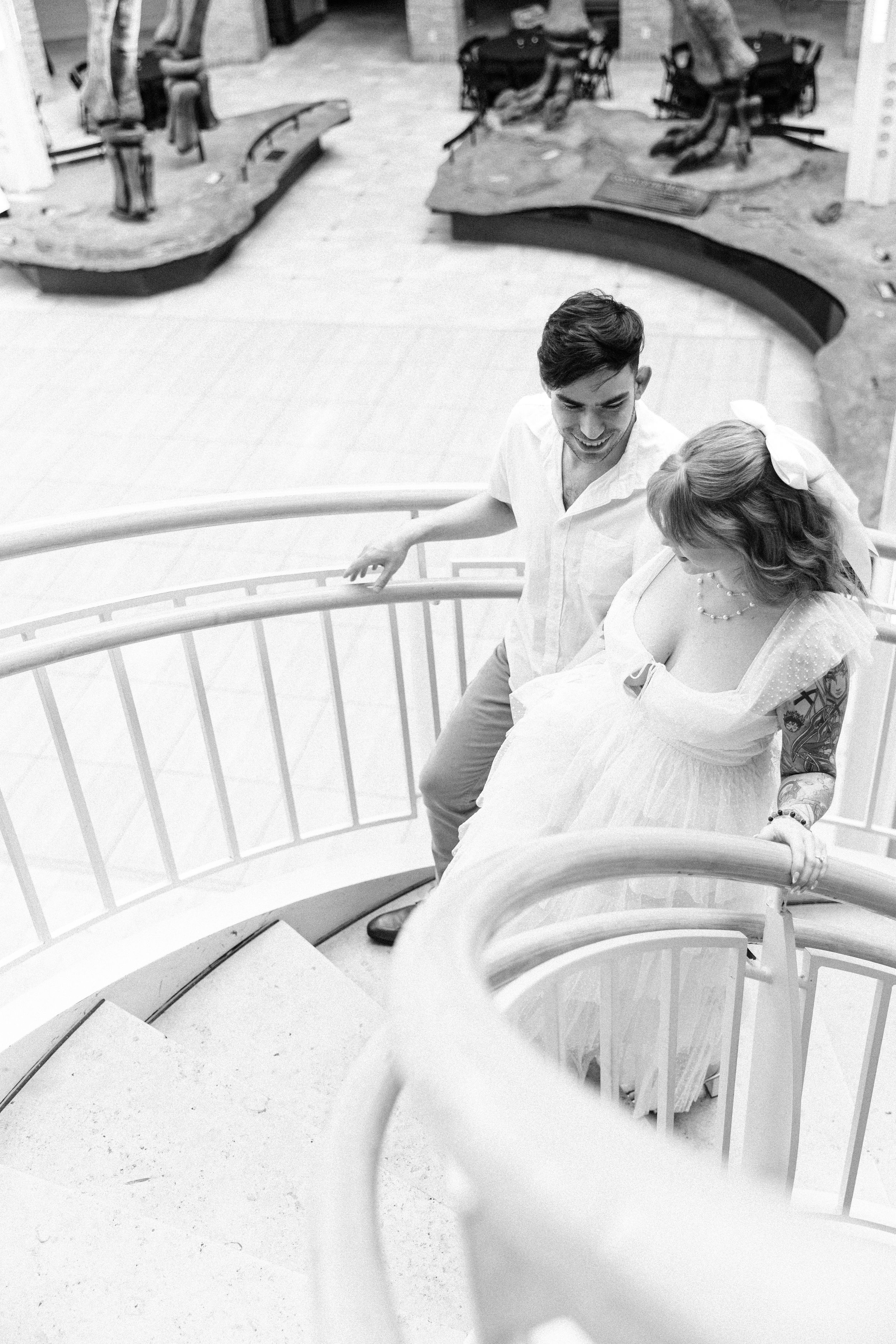 A bride in a white dress descends a curved staircase in an elegant black and white photograph.