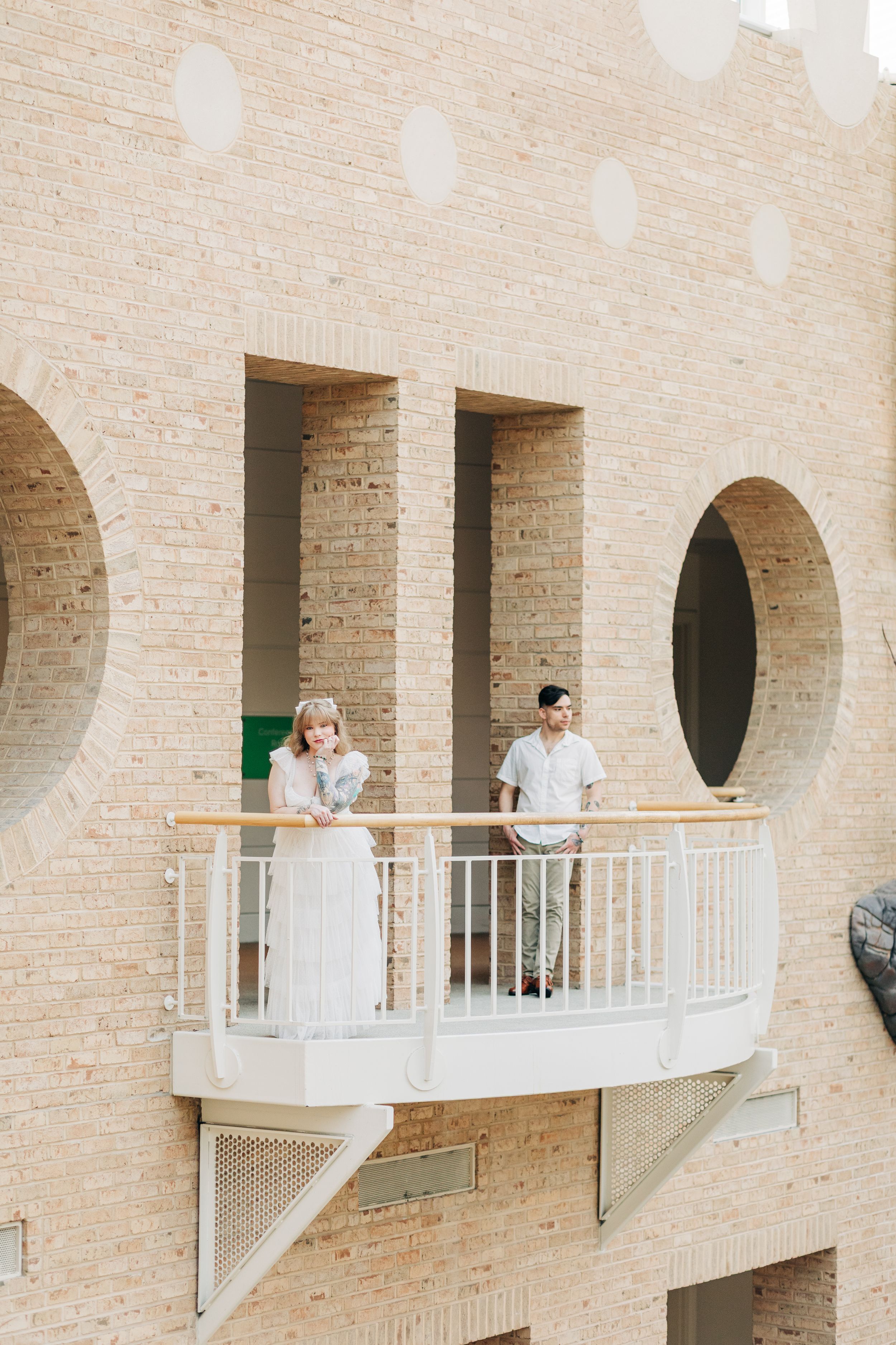 Two people stand on a modern architectural balcony with circular windows and beige brick walls in a contemporary setting.