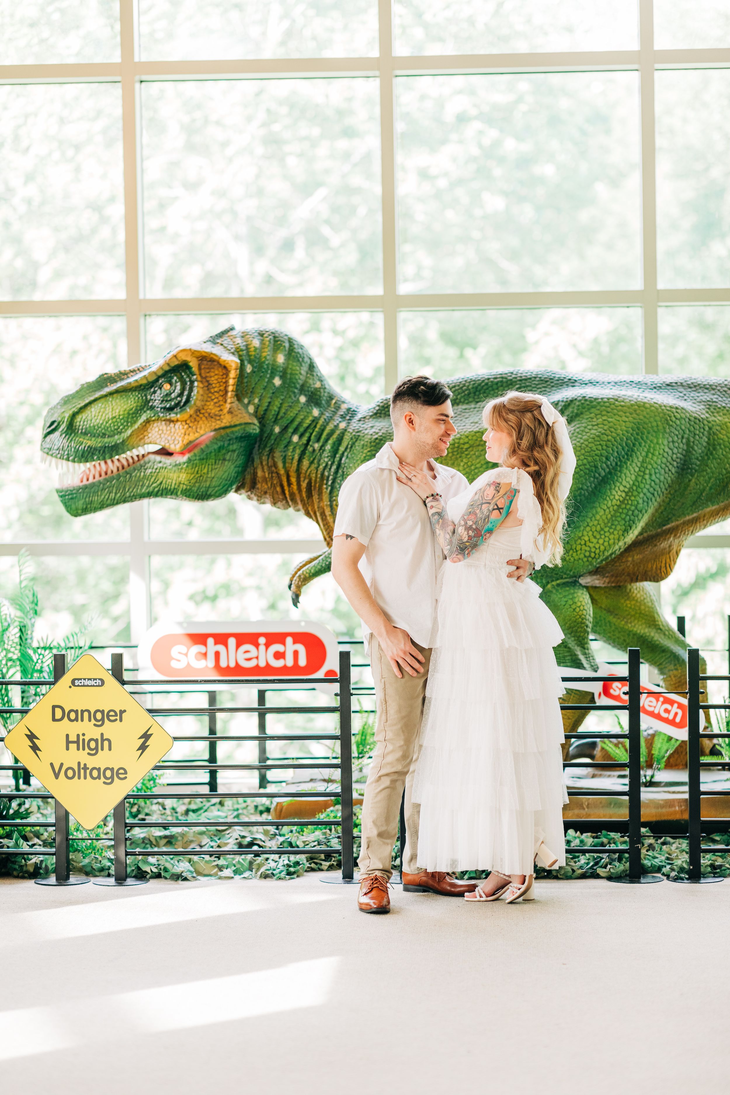 A romantic couple poses in elegant attire in front of a large green dinosaur display near a Schleich sign and window.