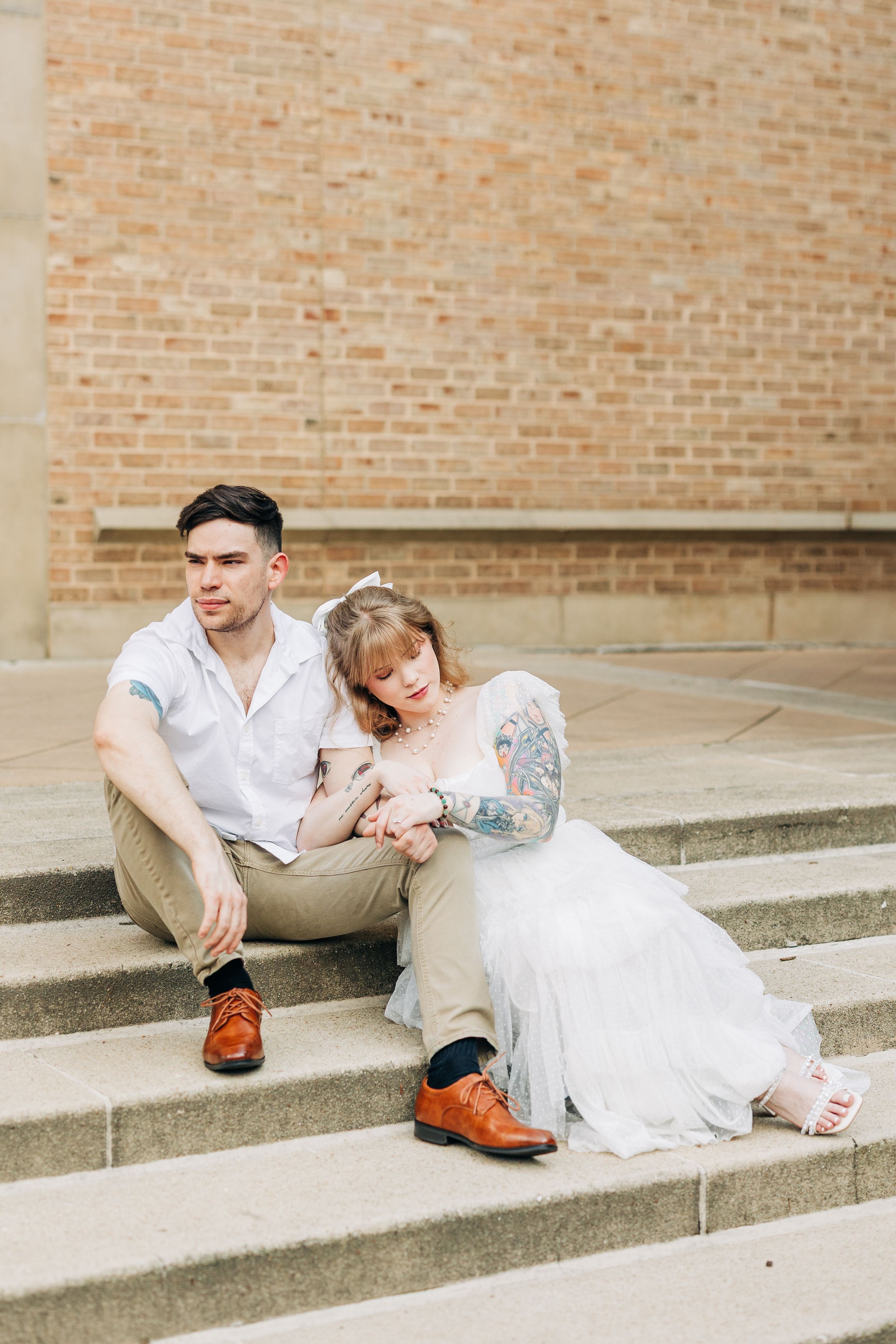 A couple in wedding attire sits relaxed on concrete steps against a brick wall backdrop.
