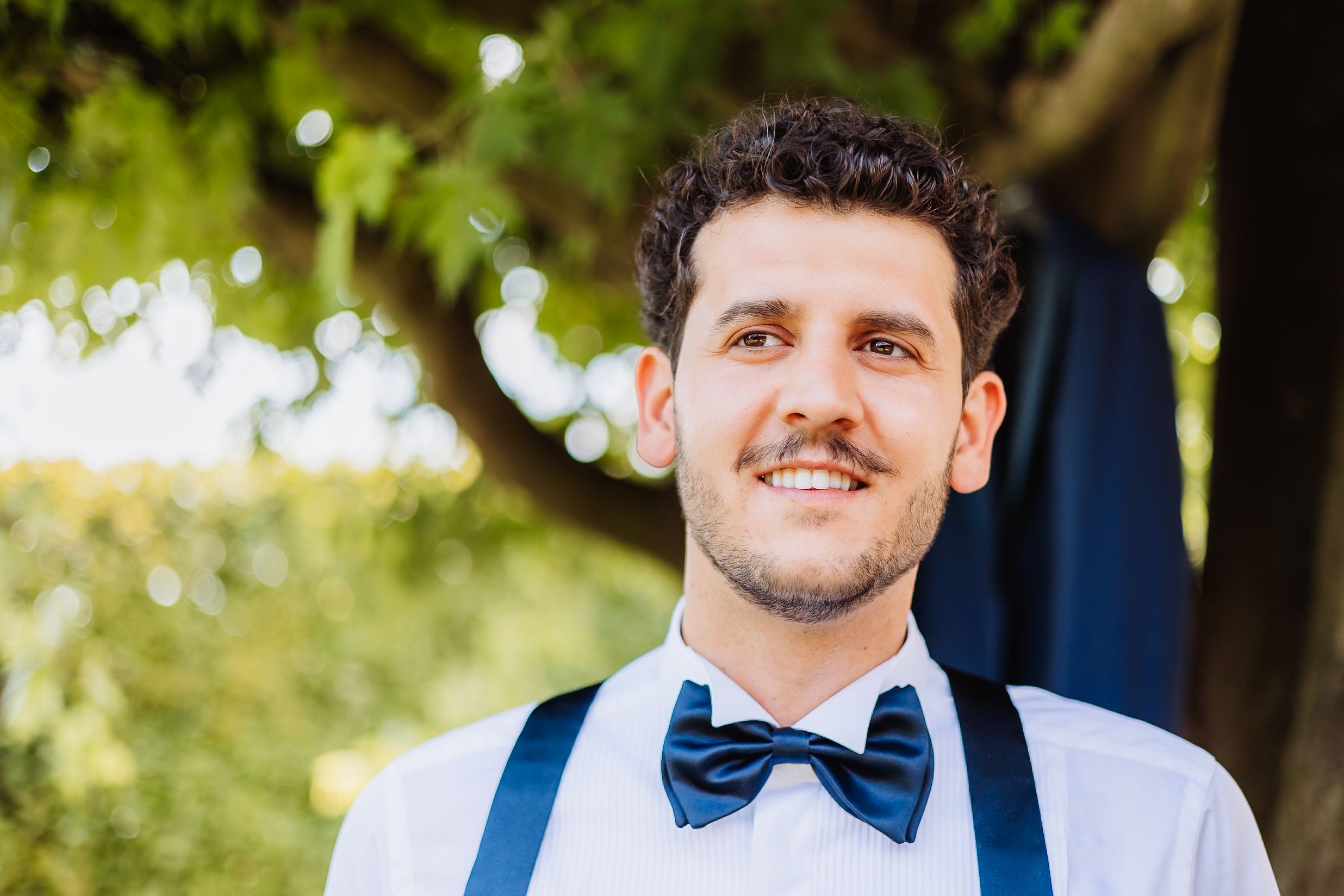 Smiling groom in white dress shirt, navy bow tie and suspenders at outdoor wedding venue with trees in background.