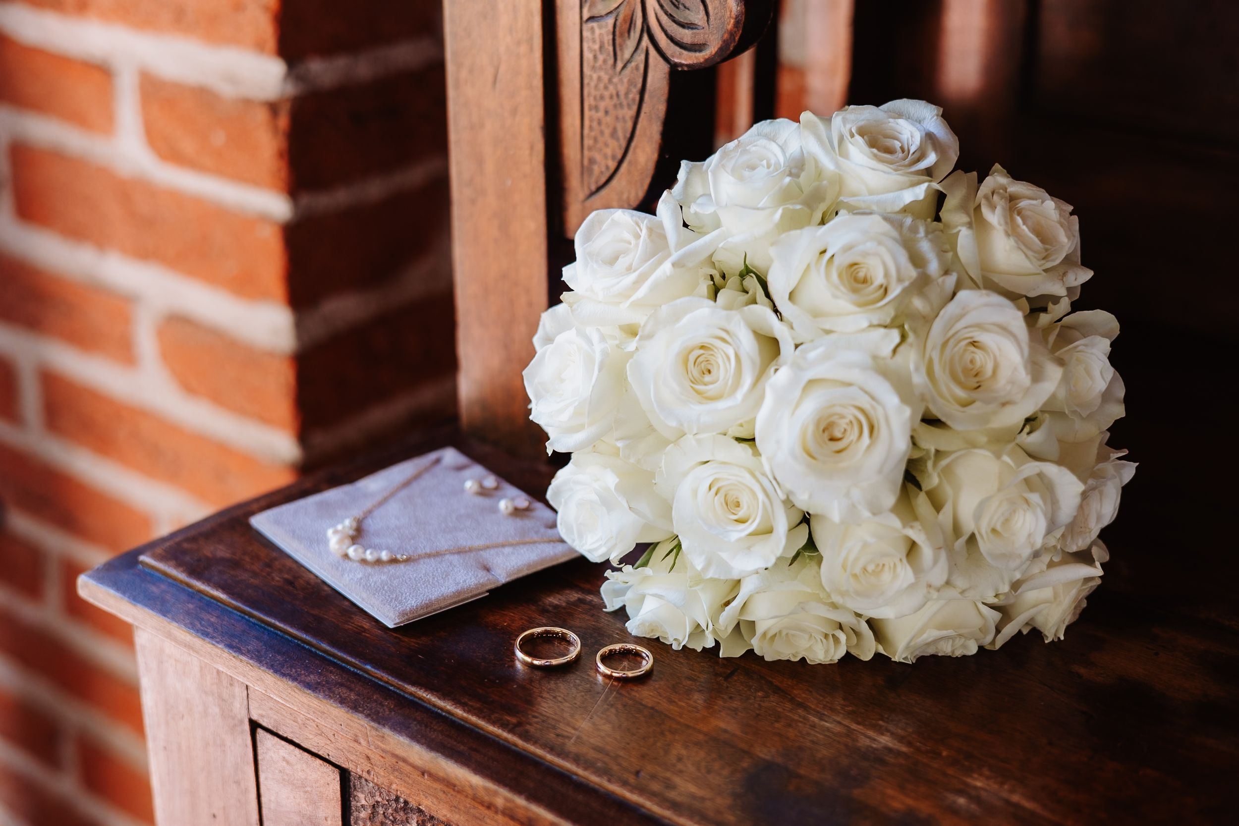 White rose bridal bouquet and wedding rings displayed on dark wooden surface against brick wall backdrop.