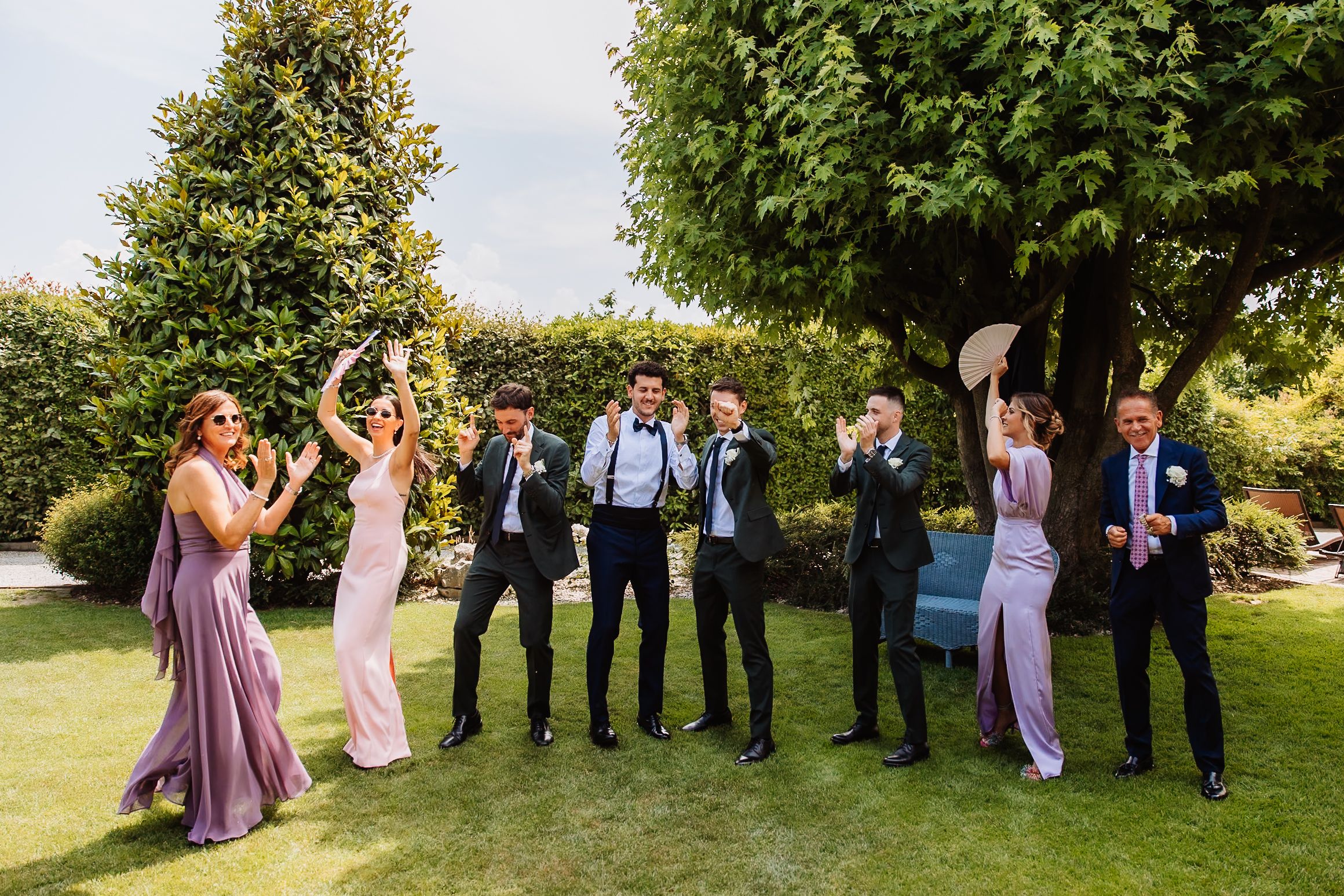 Wedding party poses for candid photos in a sunny garden setting wearing purple and grey attire.