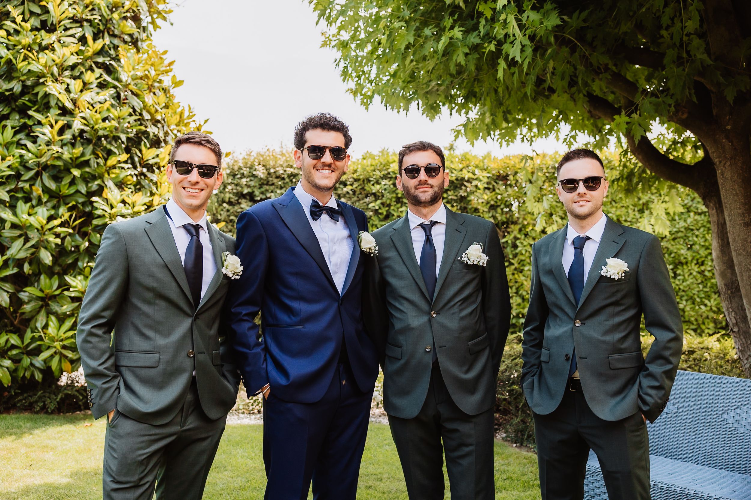 Wedding party in formal attire including navy and gray suits posing outdoors on sunny day.