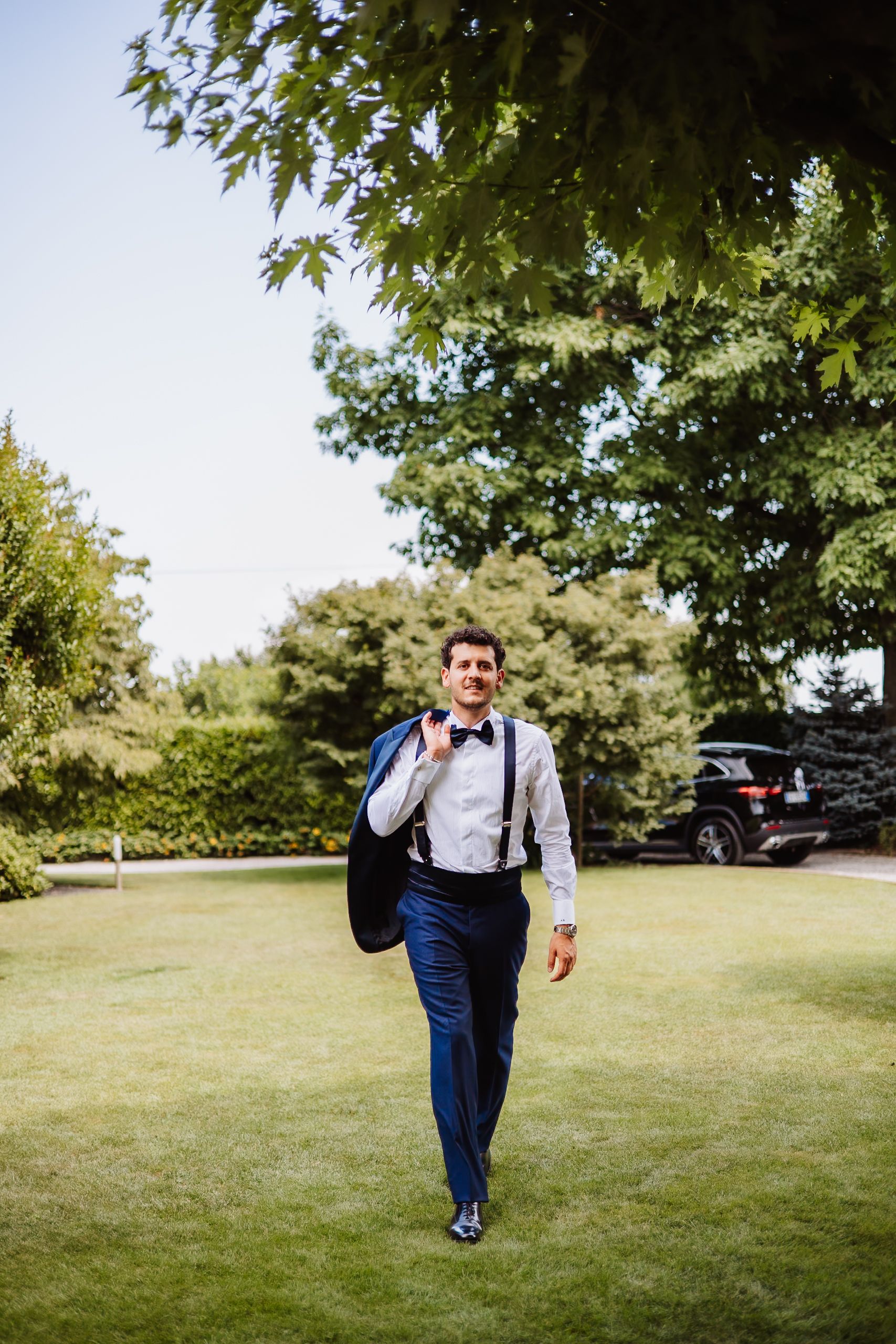 A person in formal attire with suspenders and bow tie walks across a sunny lawn with trees in the background.