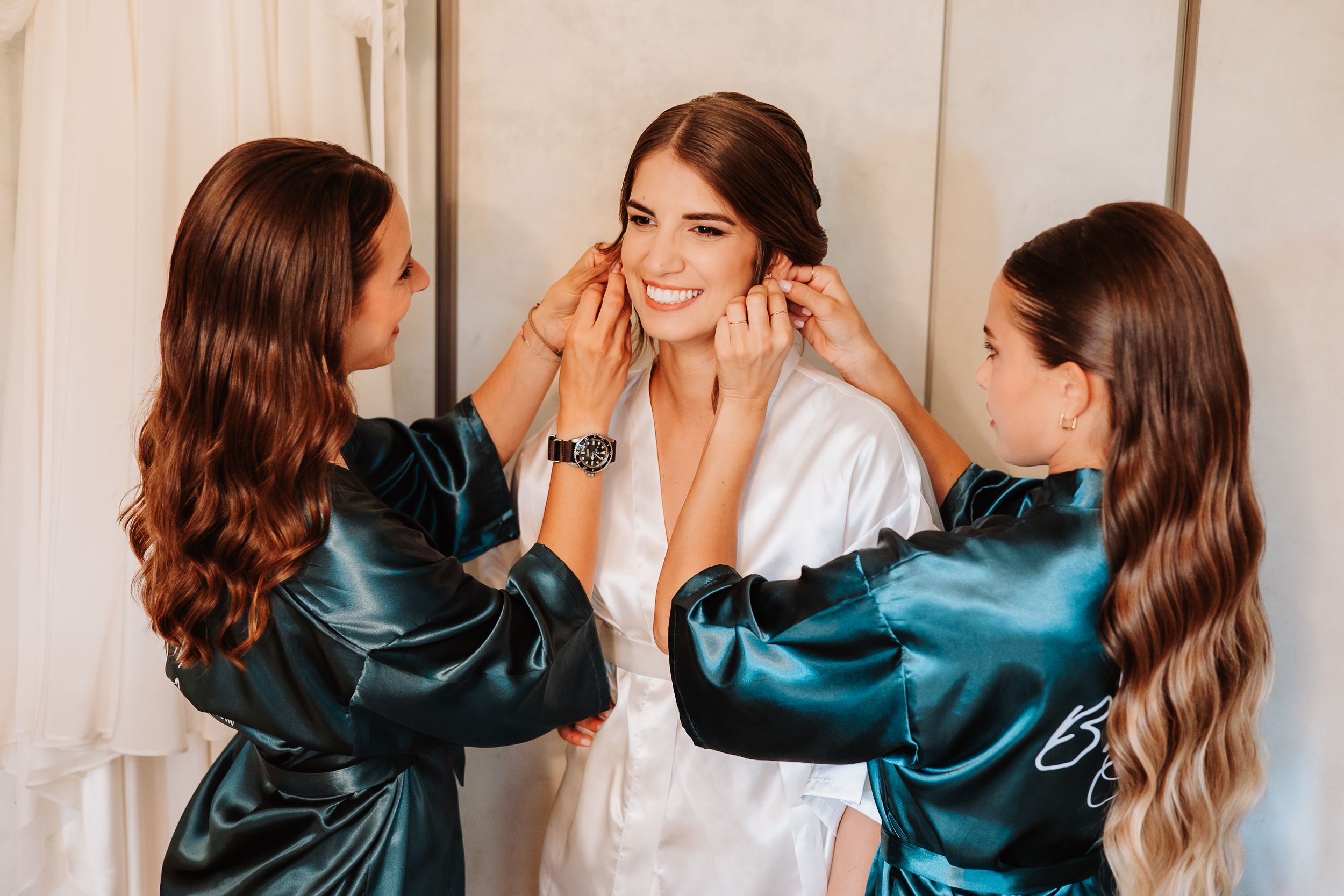 Two bridesmaids in teal silk robes help the bride prepare on her wedding day while all smile joyfully.