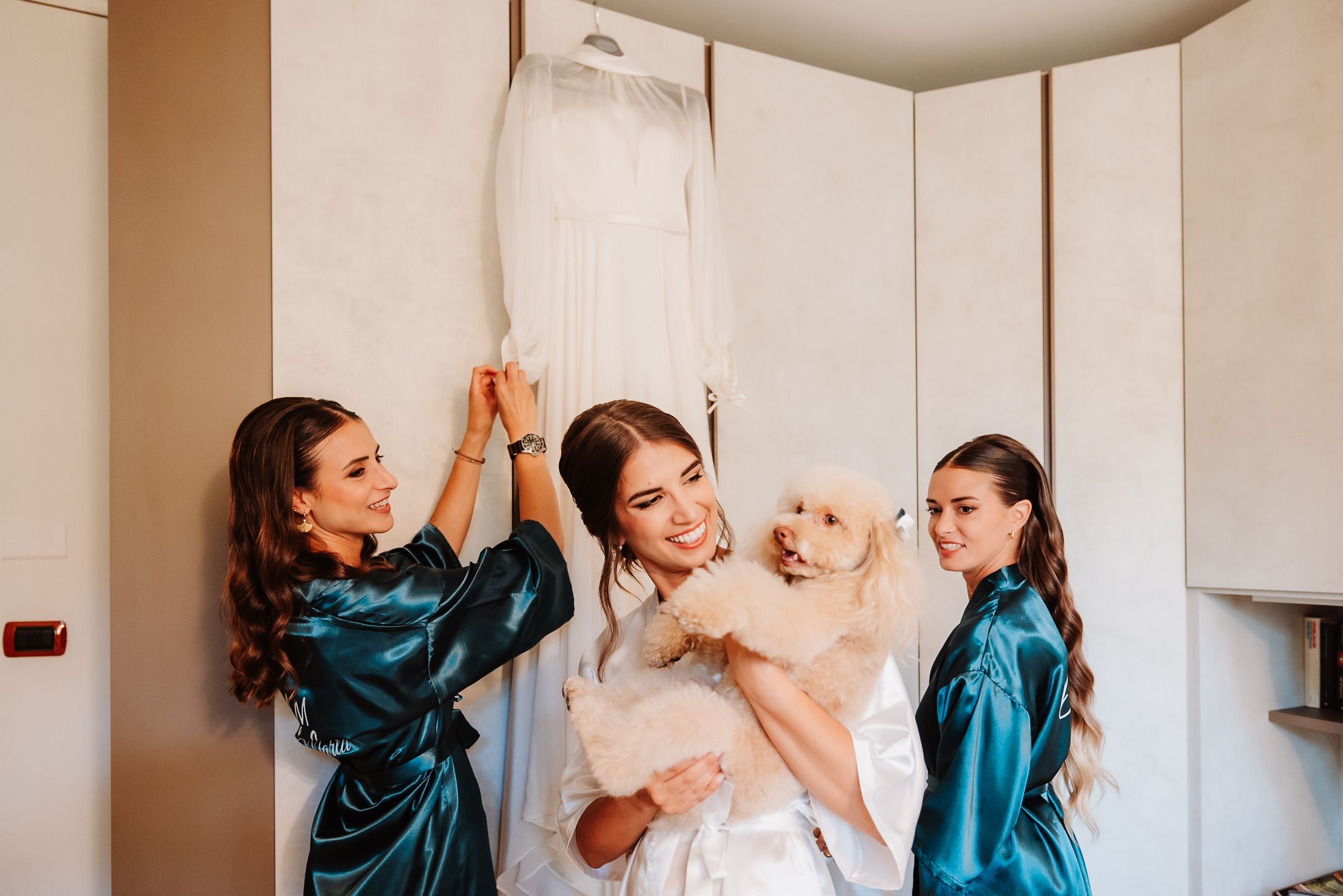 Getting ready scene in satin robes with dress hanging in wardrobe at wedding preparation.