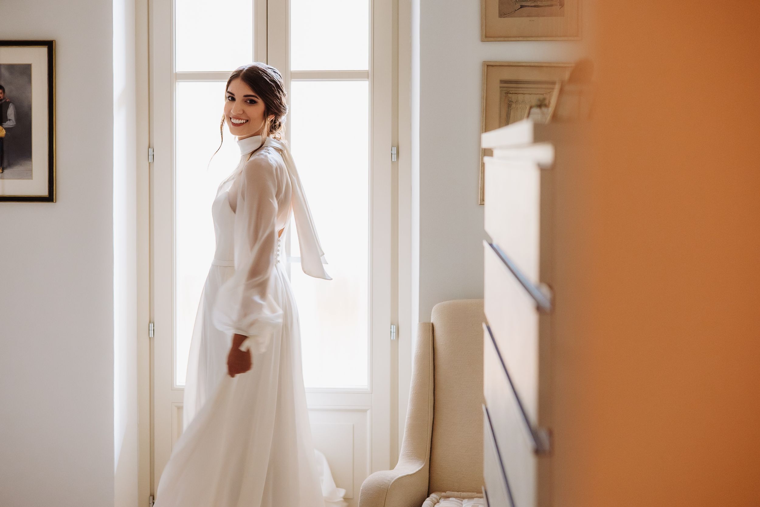 Elegant bride in long-sleeved white wedding dress stands gracefully next to sunlit window in tranquil room.