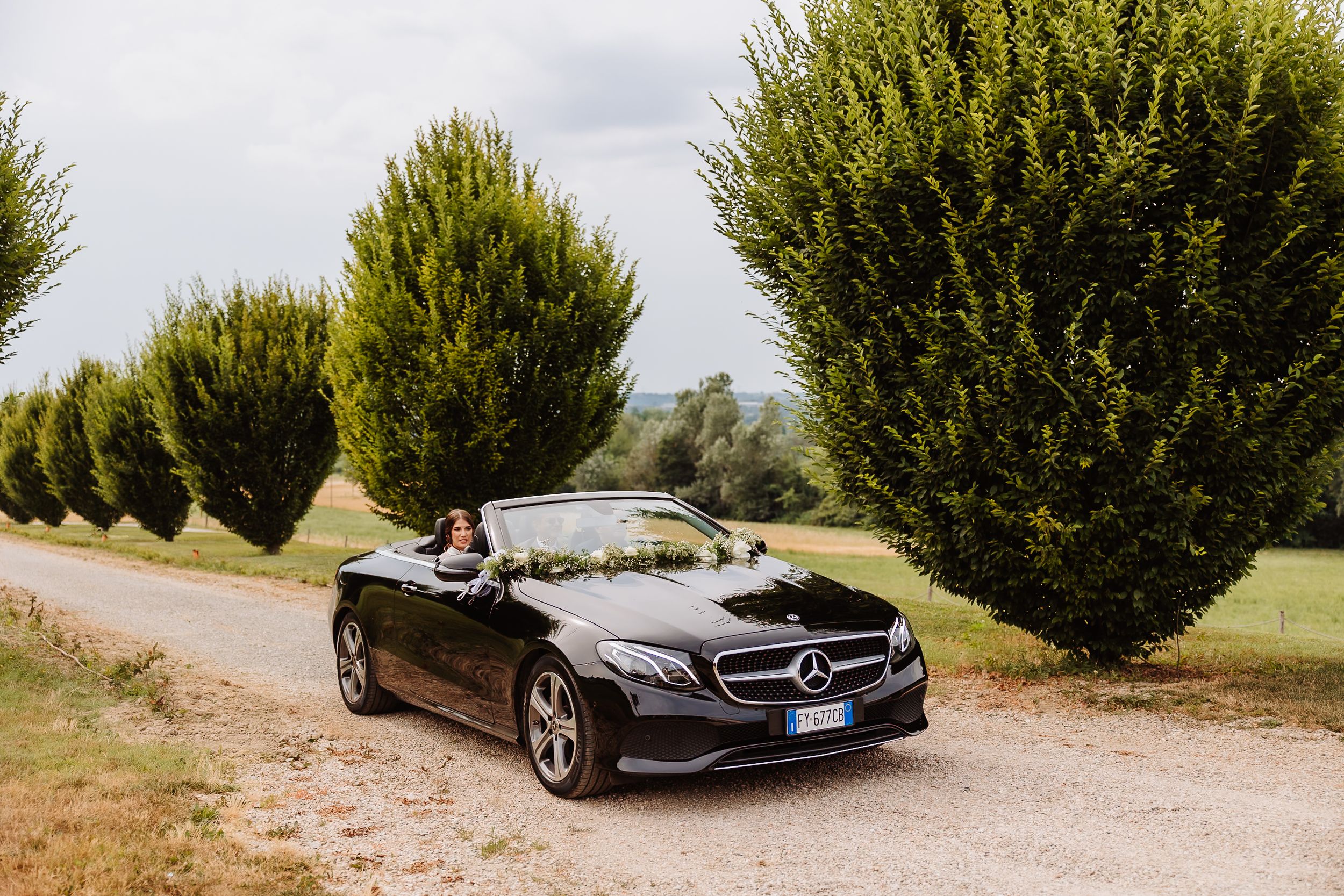 A black Mercedes-Benz convertible parked on a dirt road with tall trimmed hedges in the background.