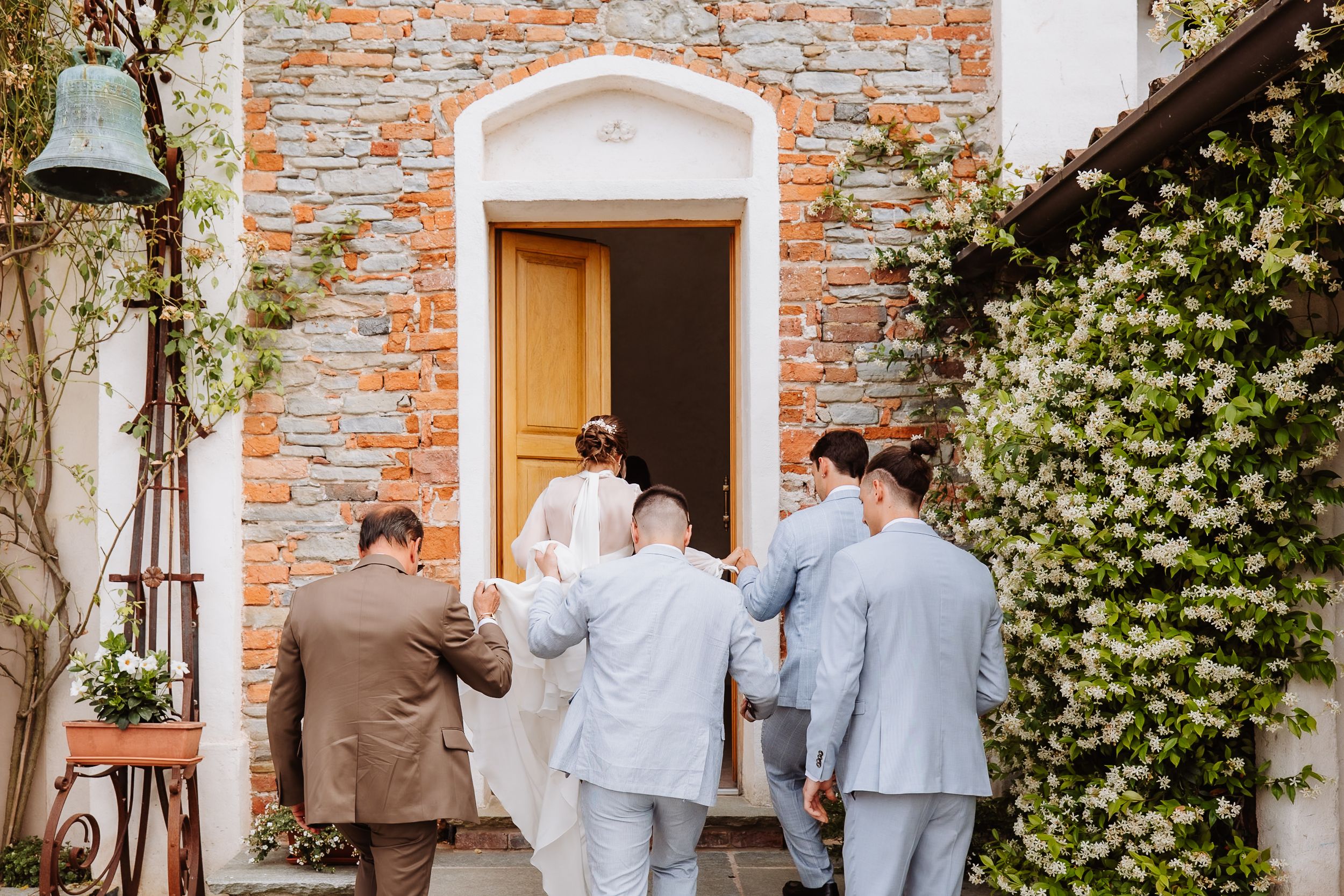 Wedding party members wearing light gray suits gather in a garden courtyard with brick walls and white trim.