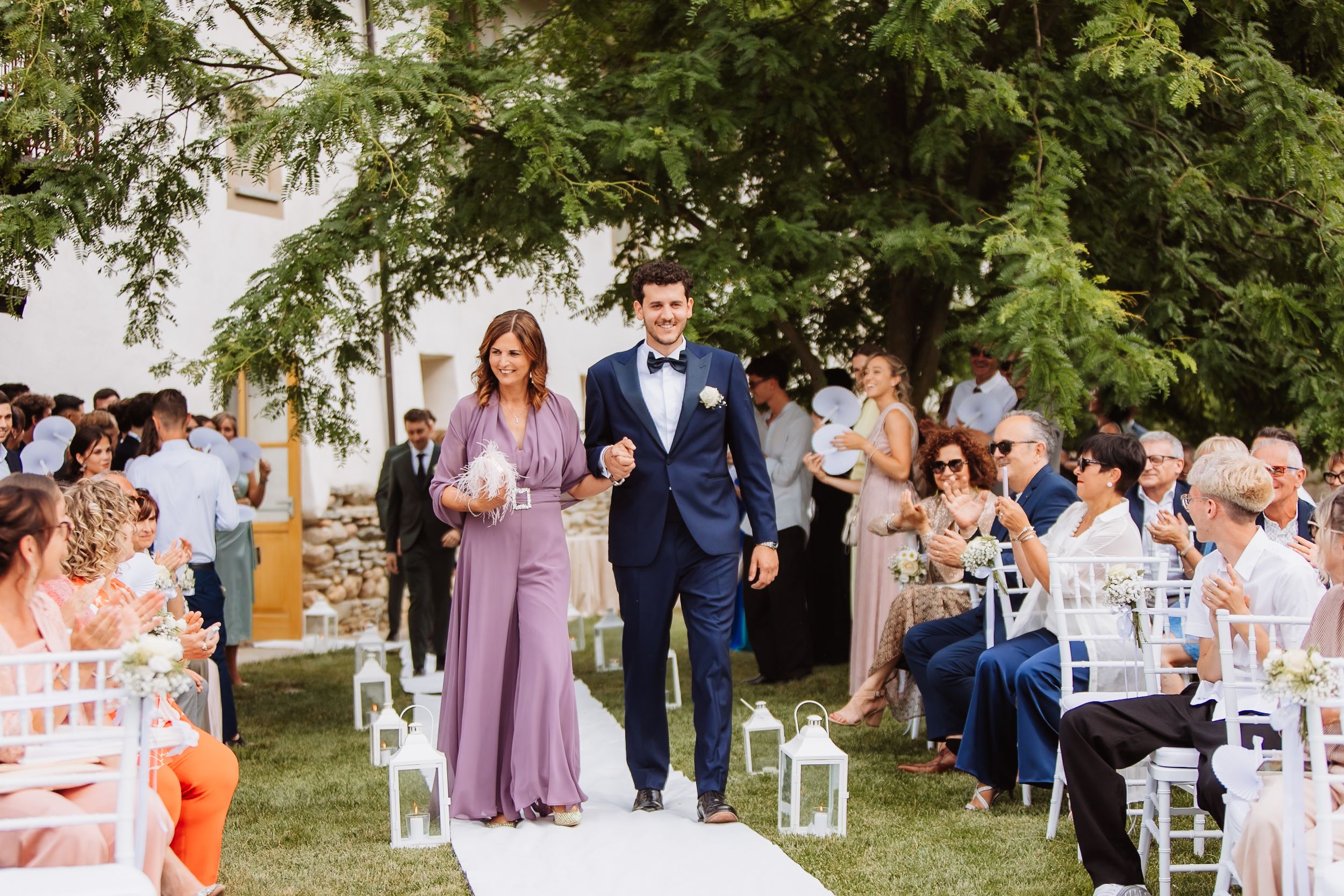Outdoor wedding ceremony on white aisle runner with seated guests on white chairs under green trees at daytime.