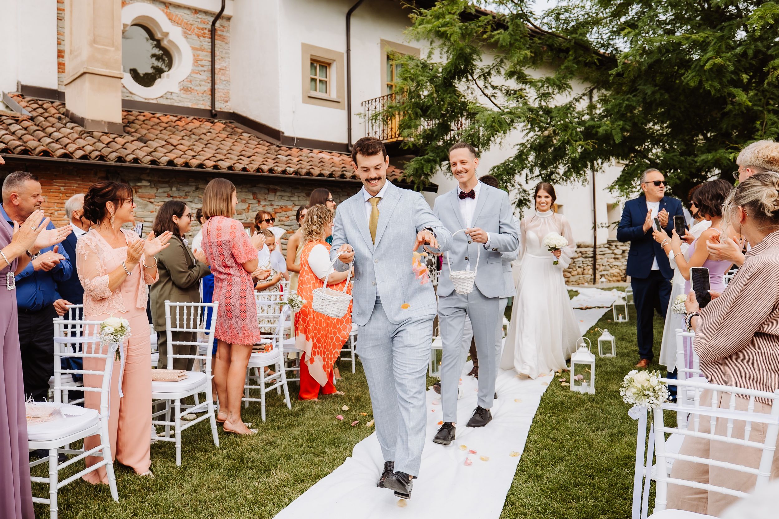 Wedding ceremony attendees celebrate as groomsmen in light colored suits walk down an outdoor aisle at a pastoral venue.