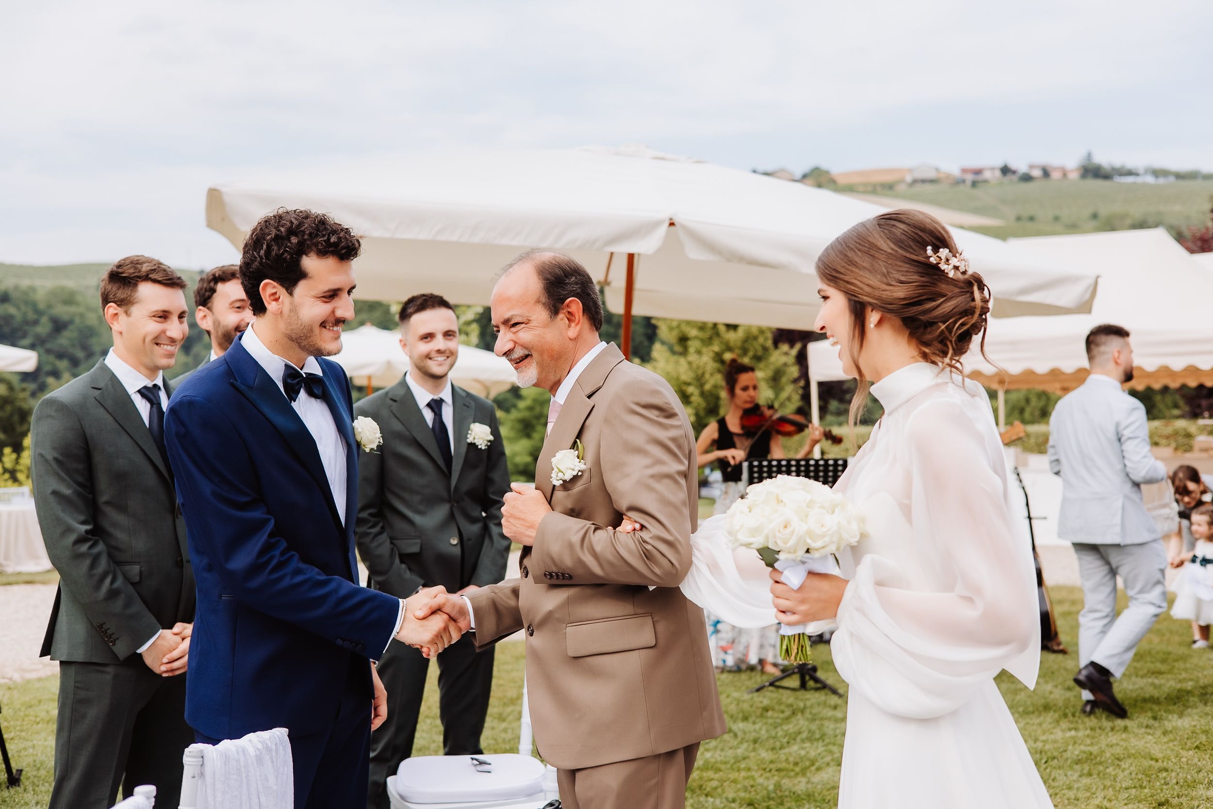 A wedding celebration takes place outdoors with guests gathered on a scenic hillside overlooking open countryside.