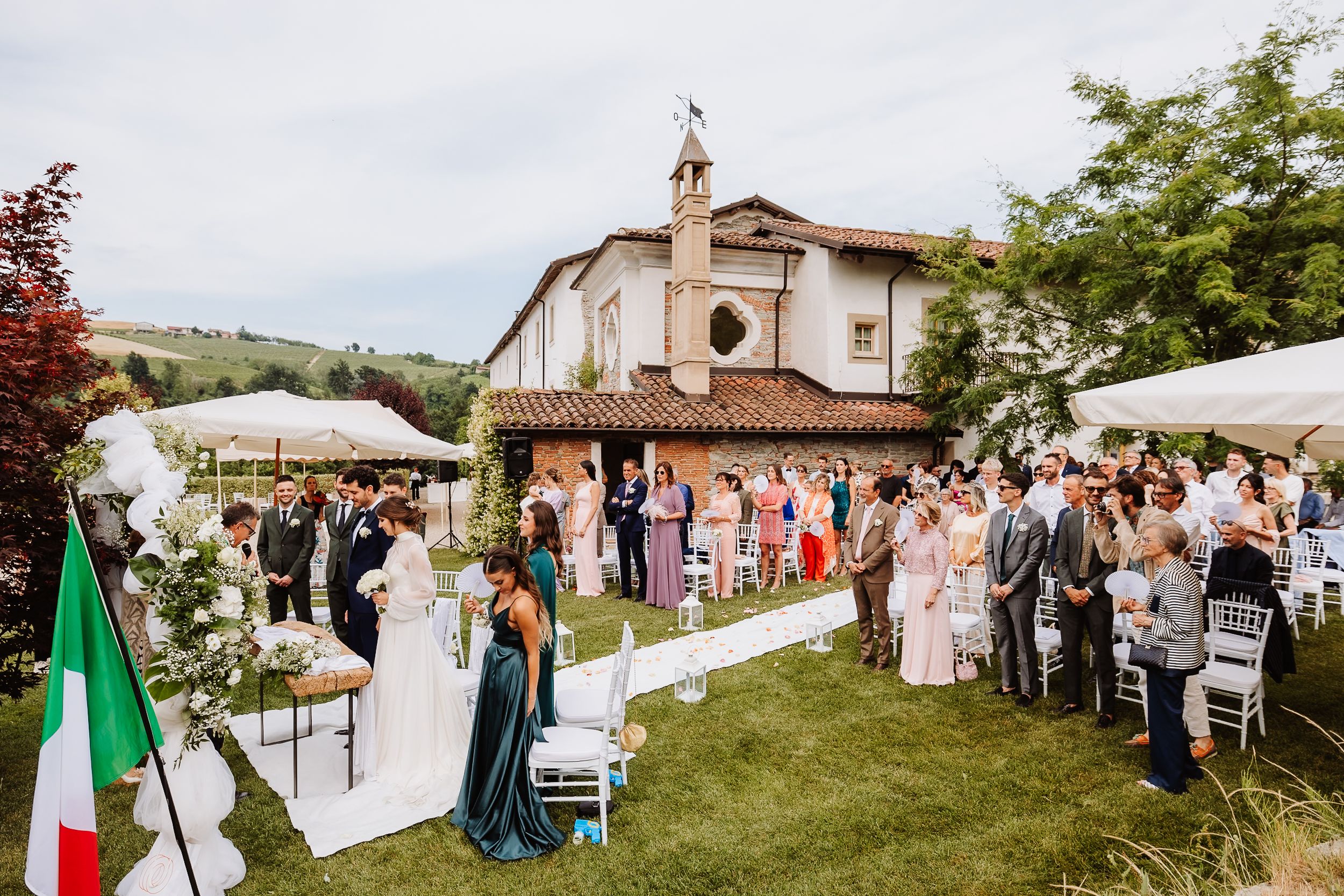 Panoramic view of an outdoor Italian countryside wedding celebration with guests gathered under a cloudy sky.