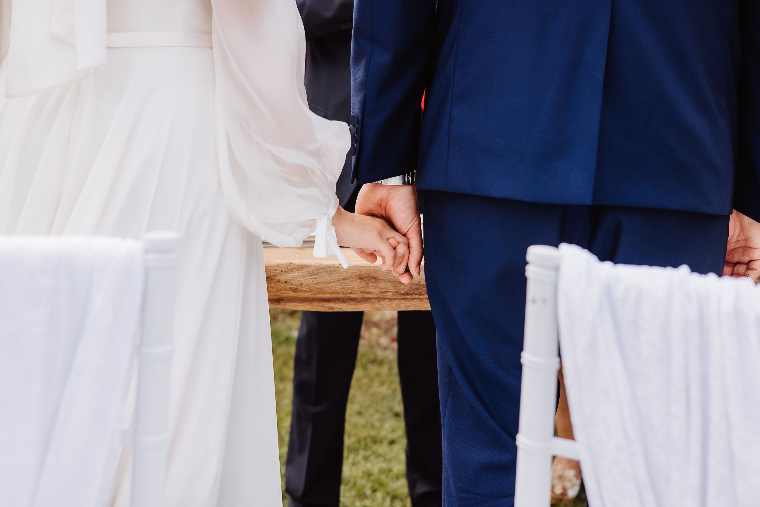 Close-up detail of clasped hands during an outdoor wedding ceremony with white chairs and blue suit attire.