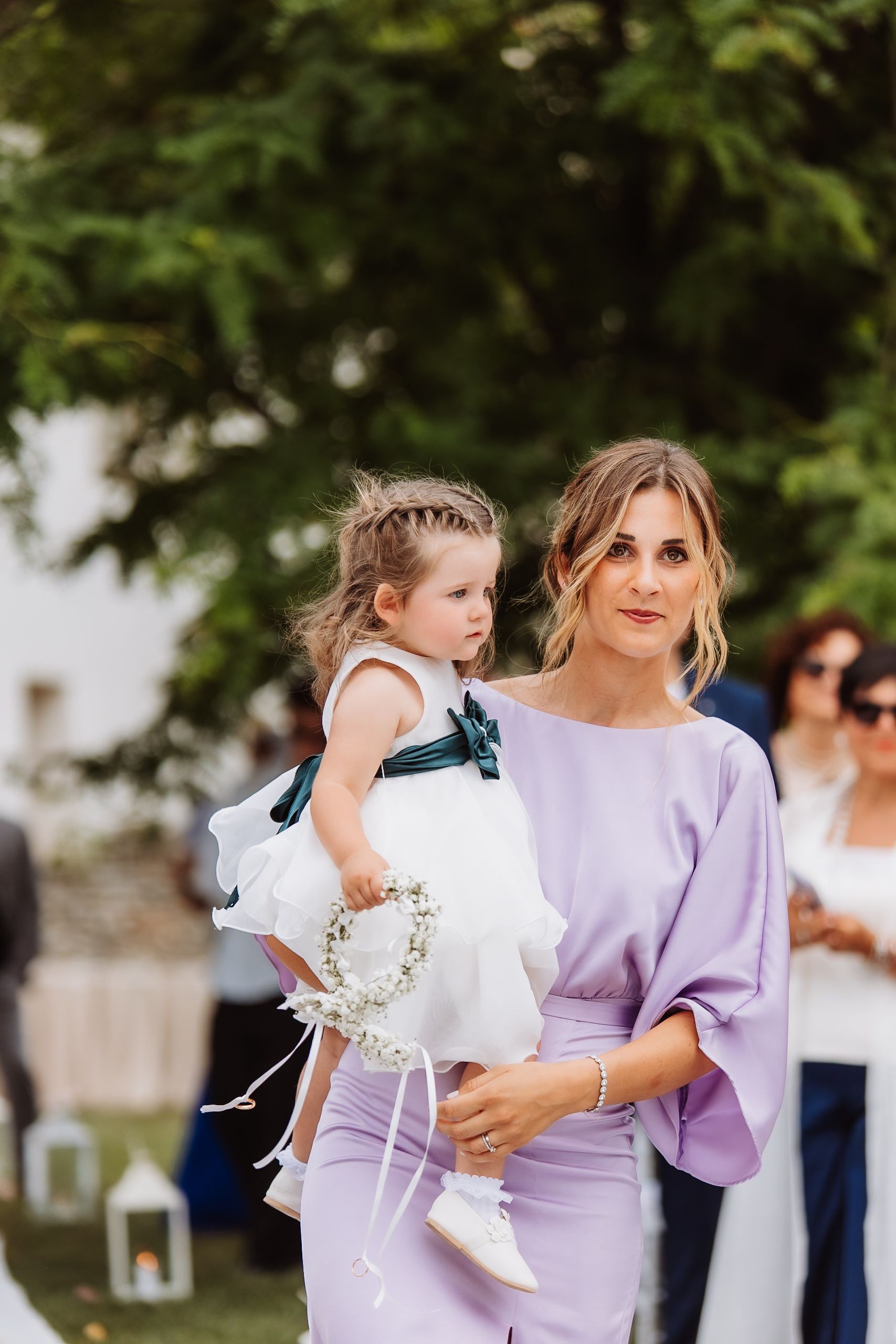 Guest in elegant purple dress holds young attendant in white dress with floral crown at outdoor wedding ceremony.