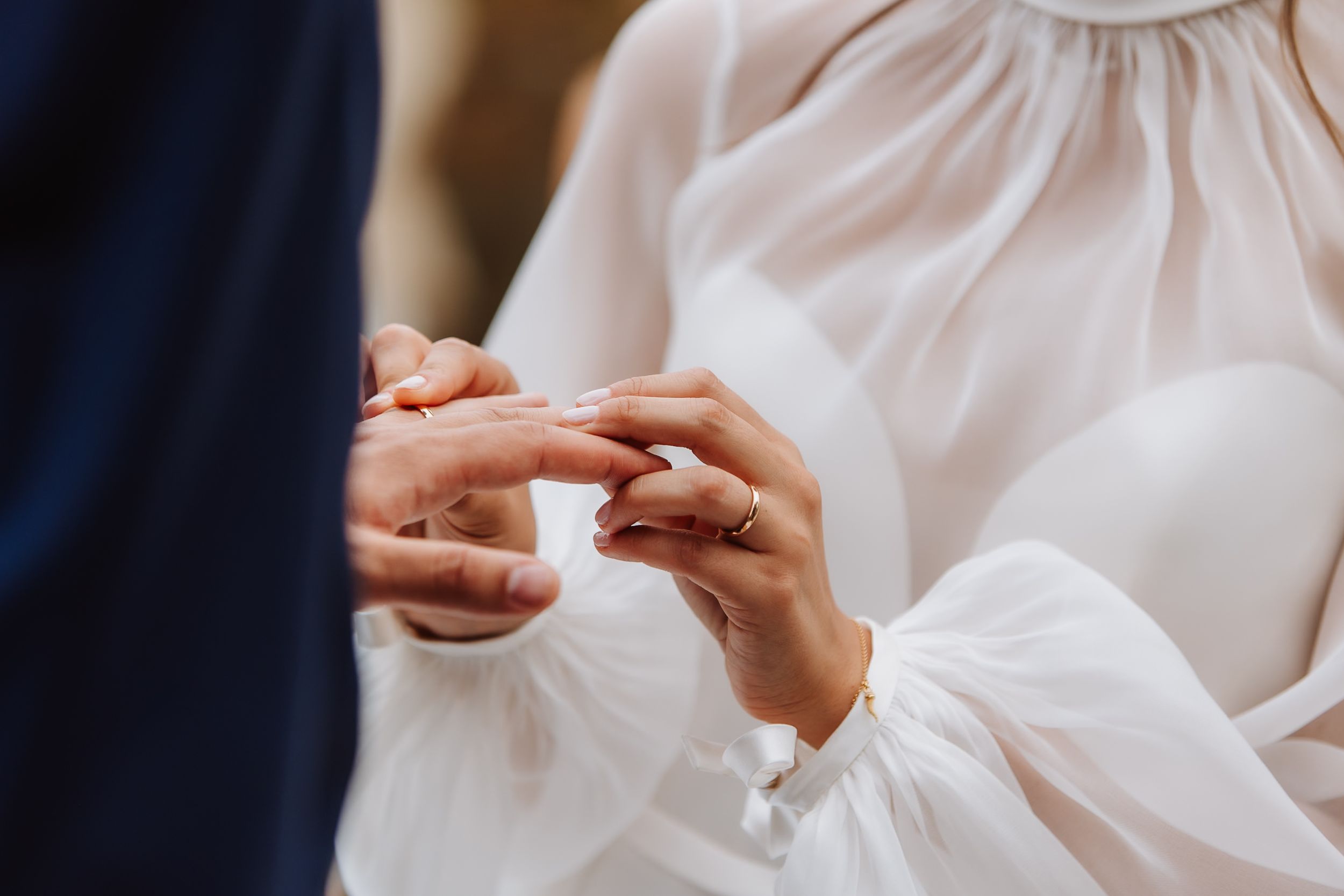 Close-up of hands exchanging rings during a wedding ceremony with white flowing fabric in the background.