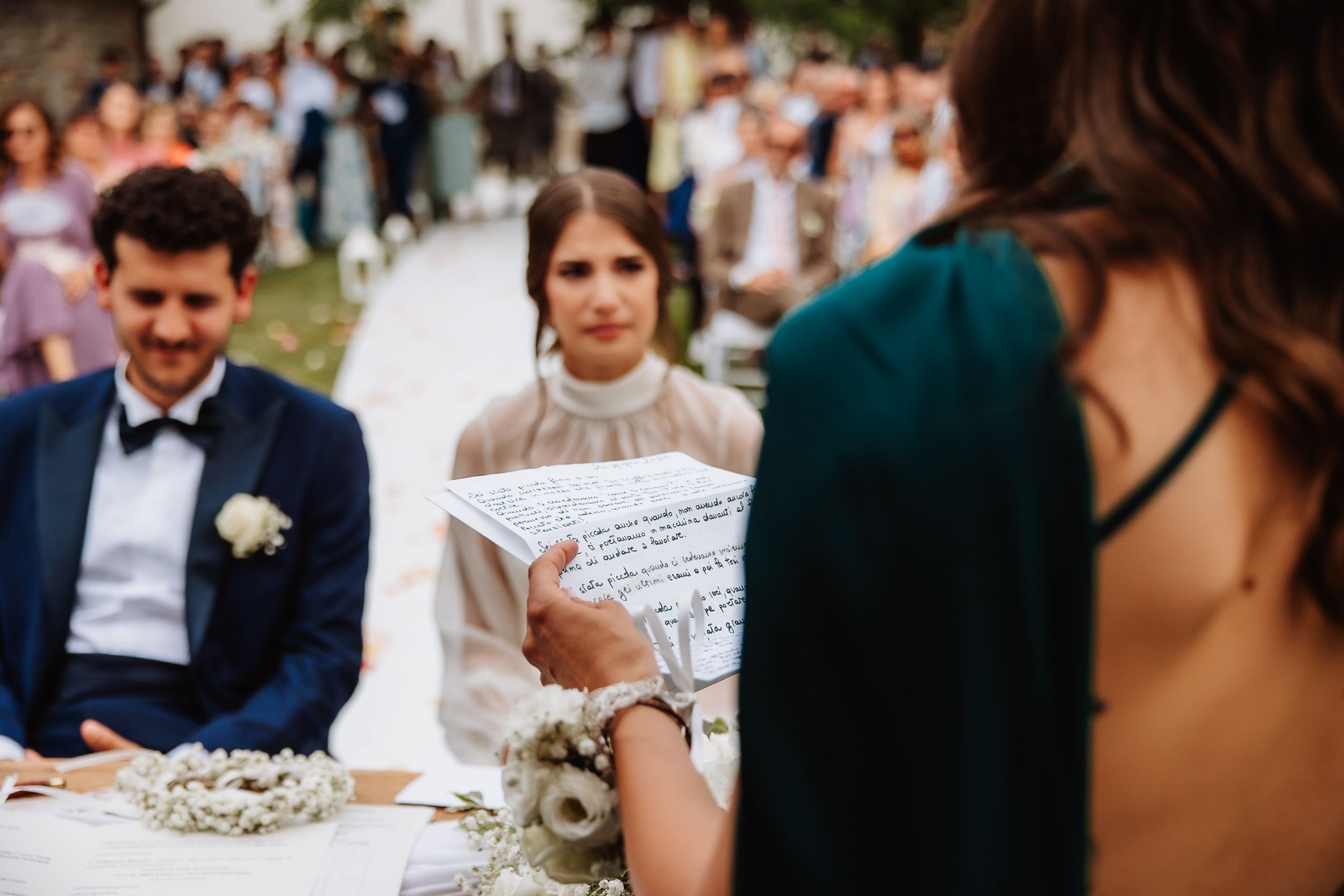 Wedding ceremony scene with guests gathered around outdoor seating while officiant reads from document.