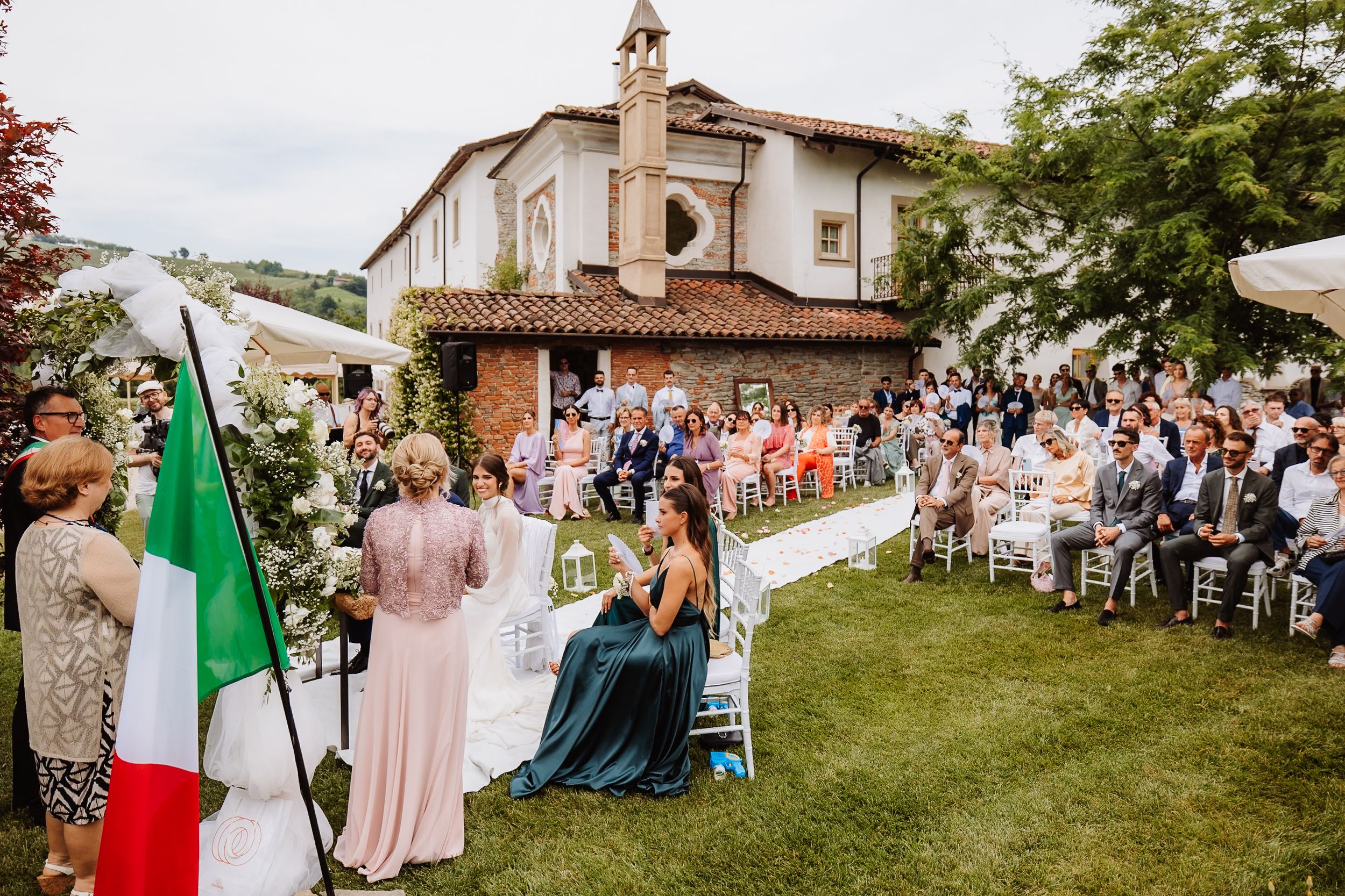 Outdoor wedding ceremony setup in a garden with white chairs and a natural backdrop on a sunny day in Italy.