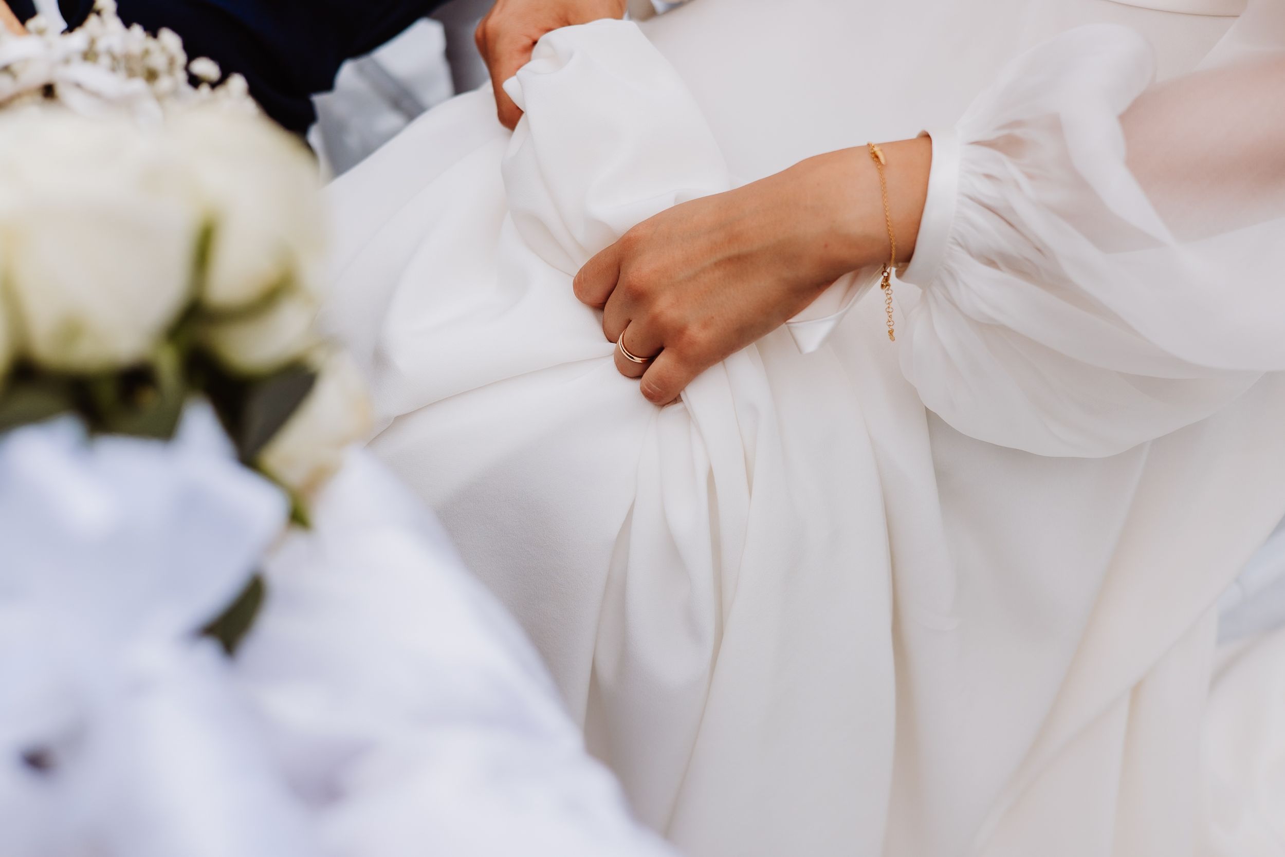 Close up of hands holding delicate white fabric details of a wedding dress with soft floral decorations nearby.