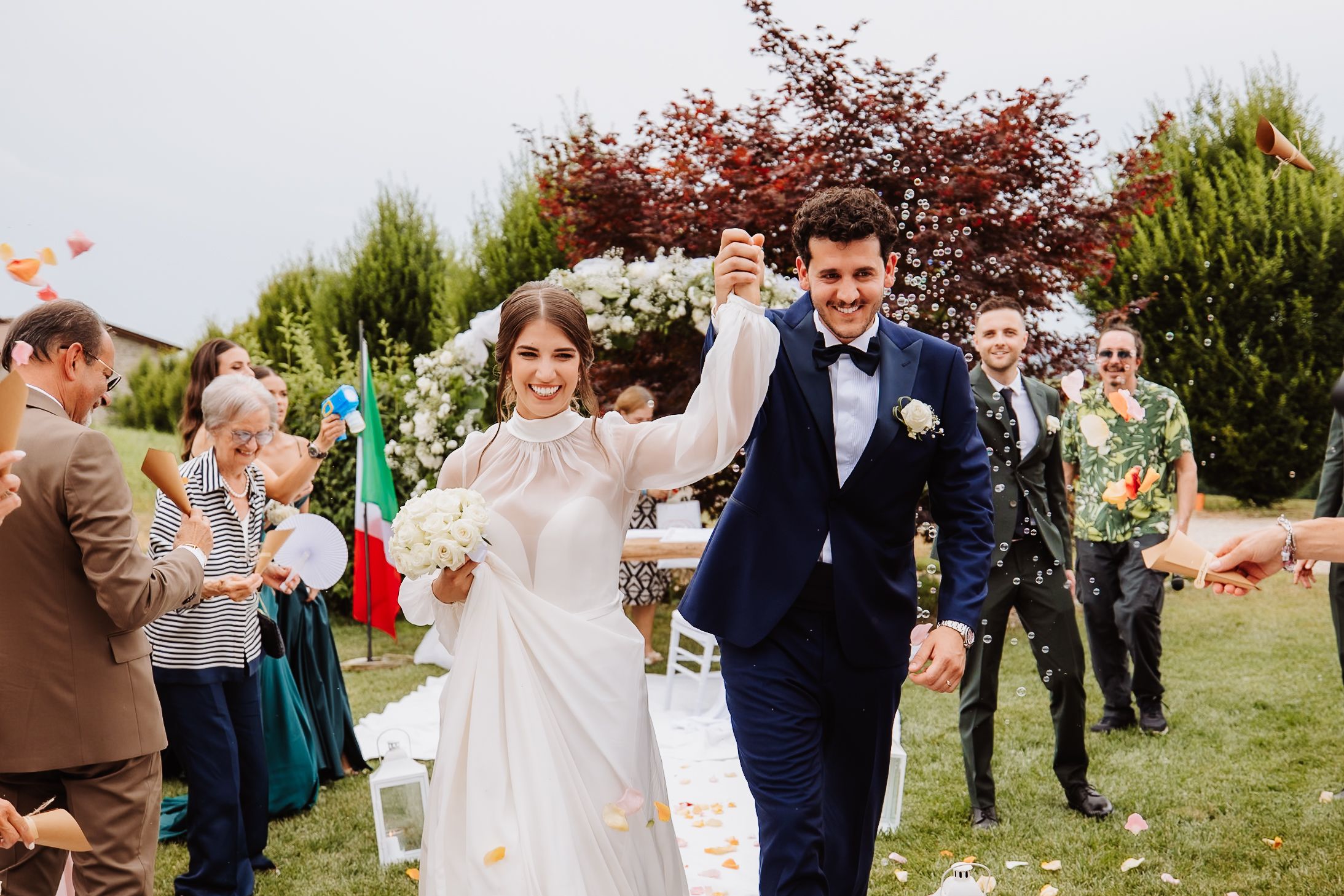 Newlyweds joyfully exit their outdoor garden ceremony while guests shower them with flower petals and confetti.