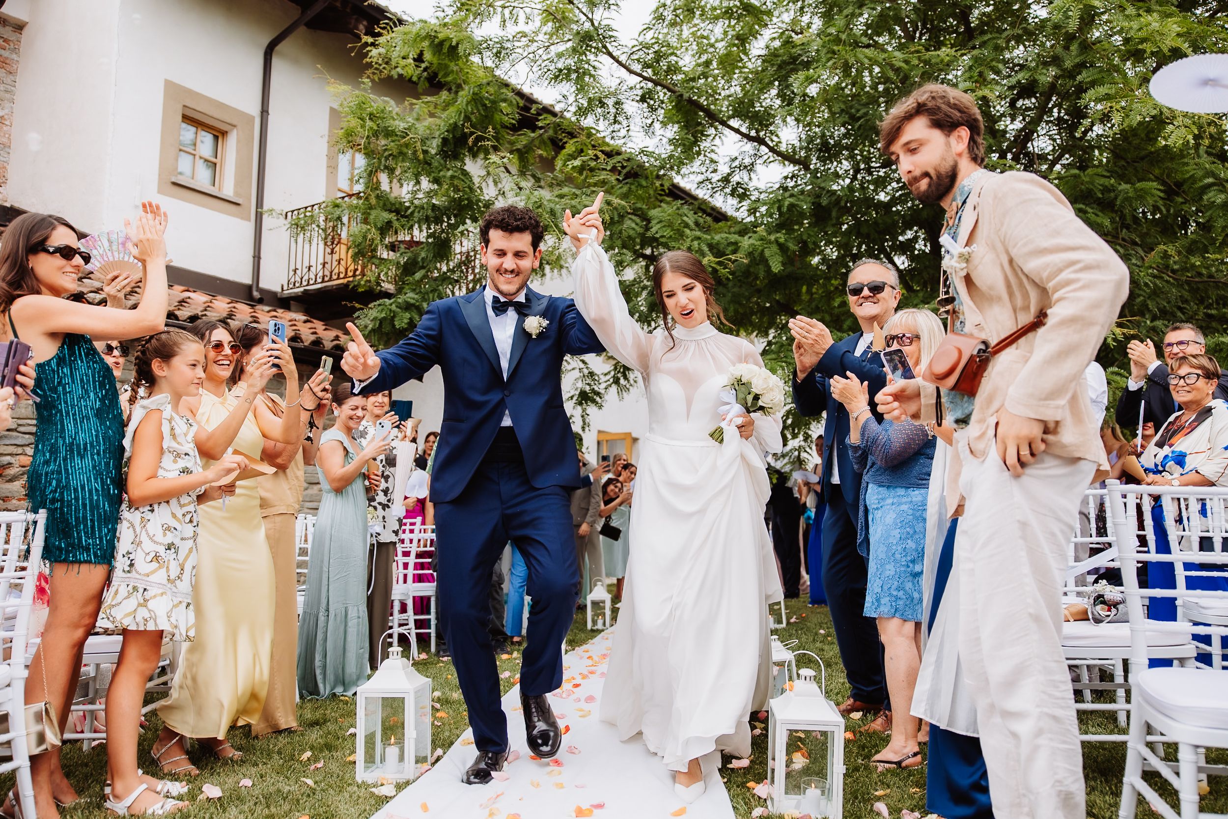 Wedding guests celebrate joyfully as couple walks down garden aisle decorated with white lanterns at outdoor ceremony.