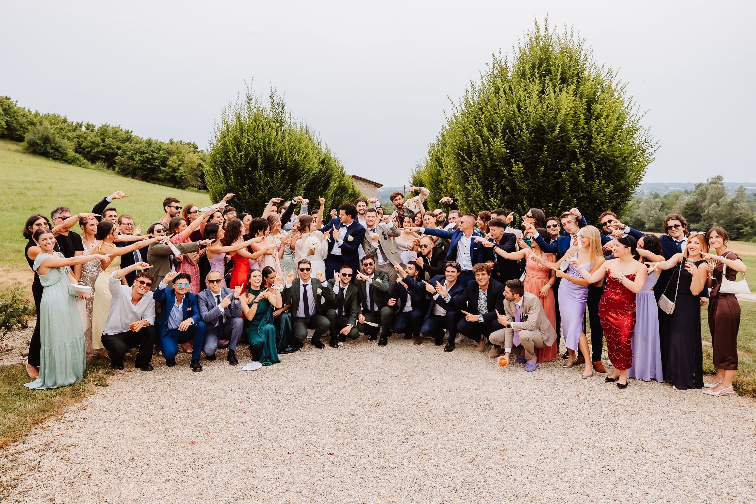 A large wedding party poses together on a gravel path surrounded by green hills on a sunny day.
