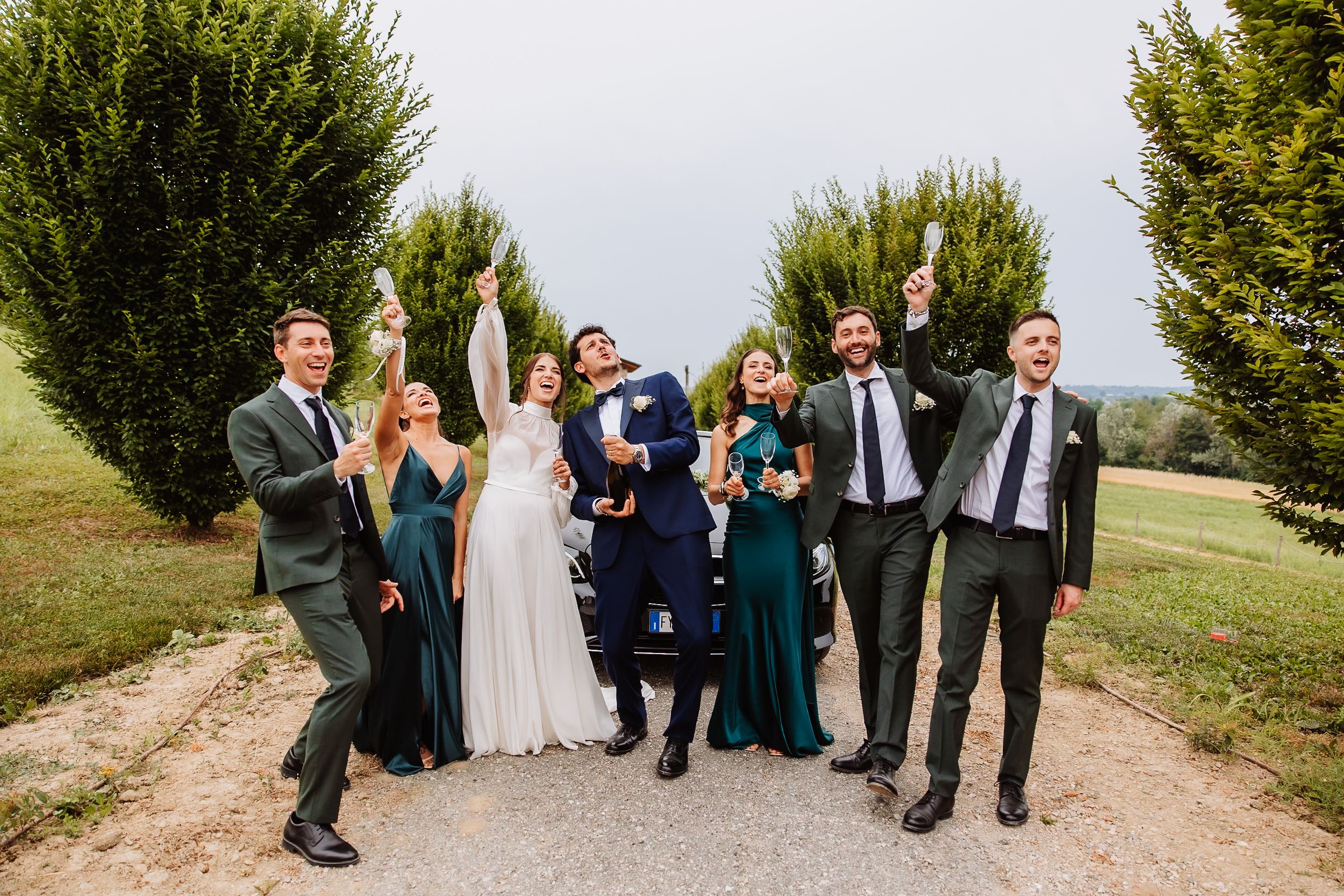 Wedding party celebrating and cheering together on a garden path surrounded by evergreen trees.