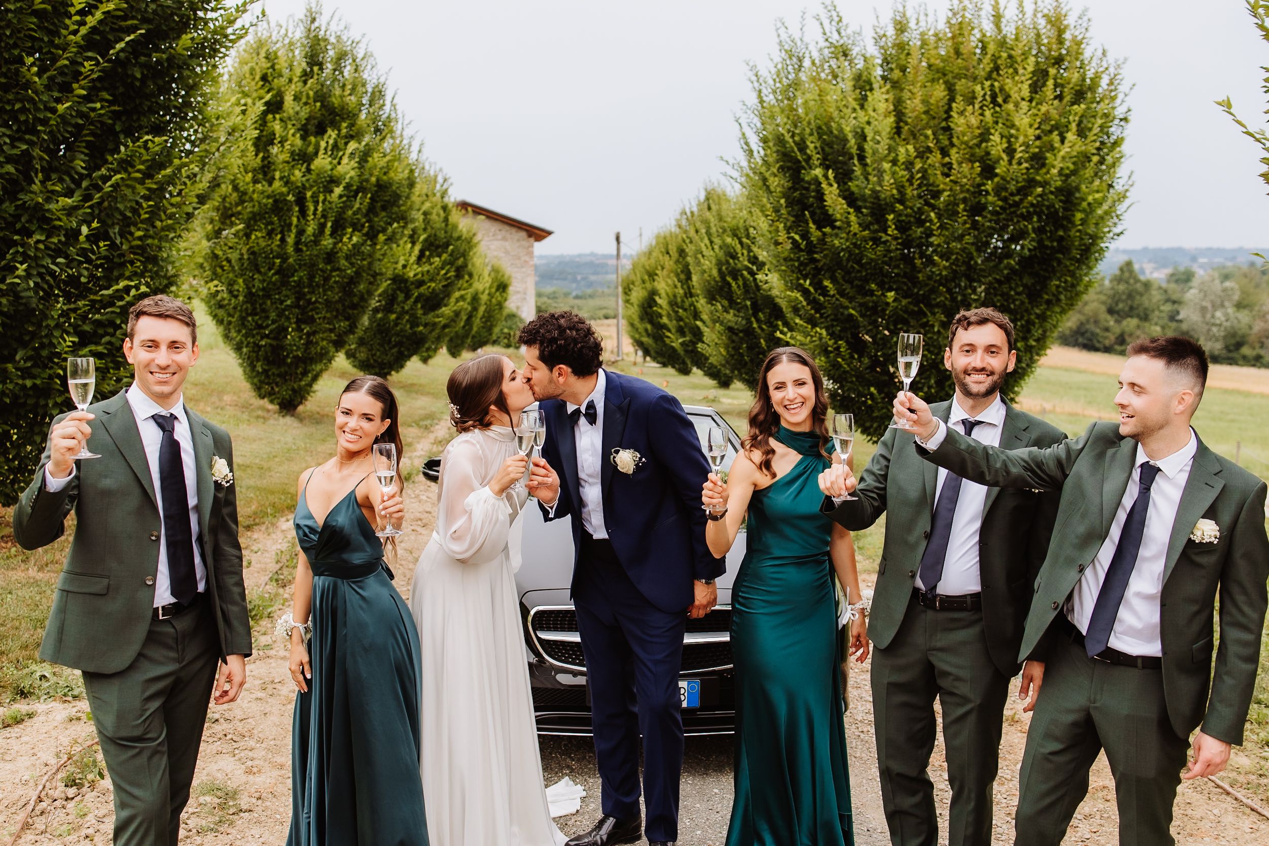Wedding party laughs together on a hillside with green trees and mountain views in the background.