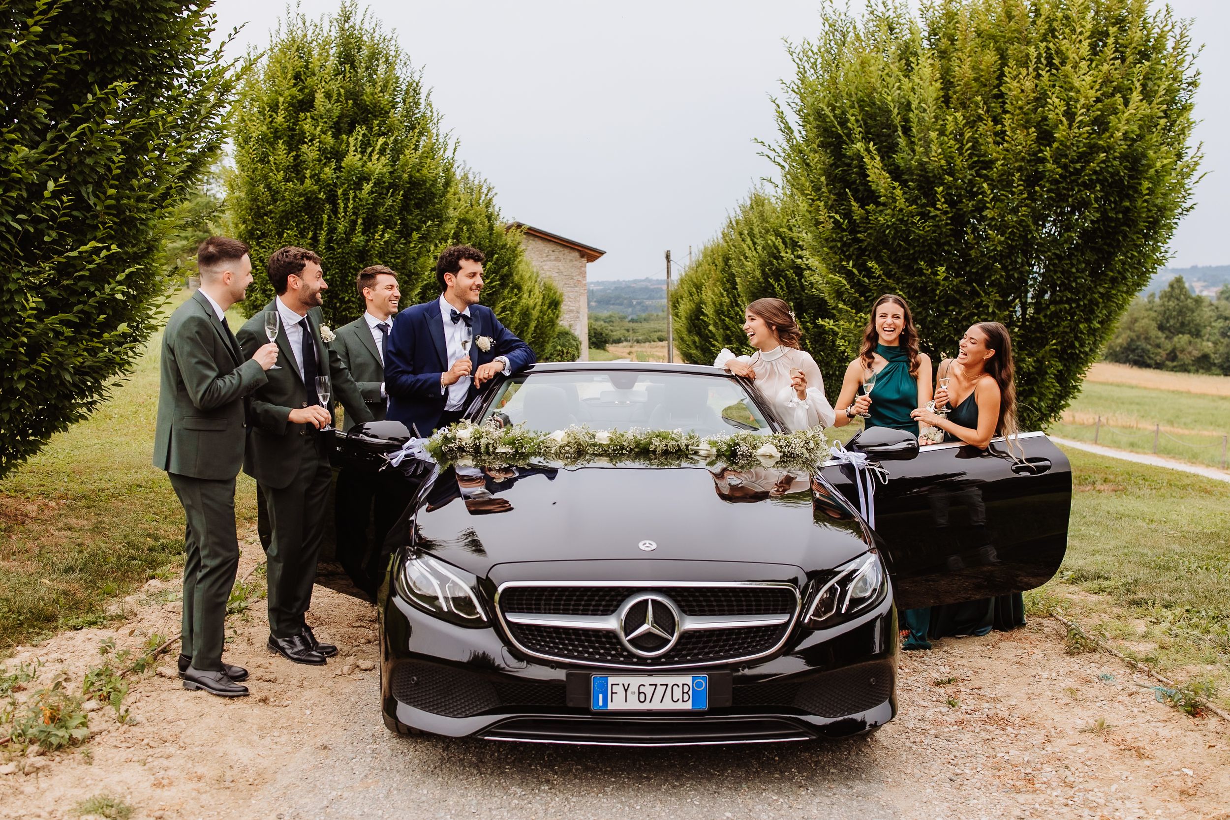 A group gathers around a black Mercedes-Benz on a country road with cypress trees in the background.