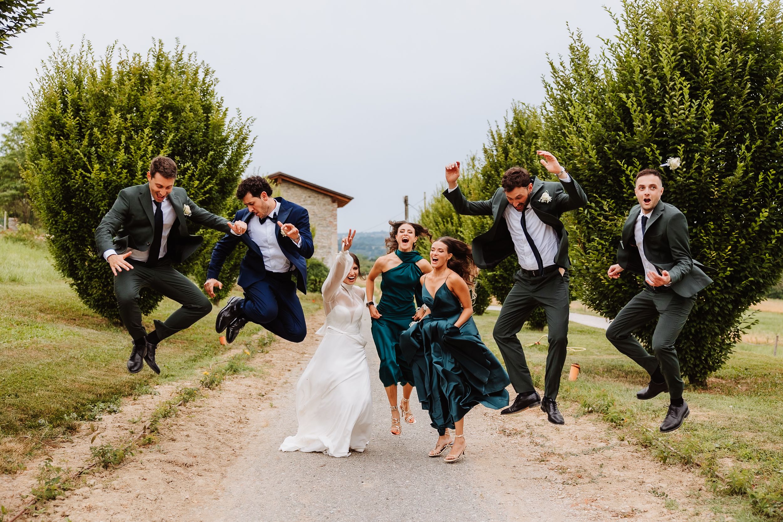 Wedding party jumps joyfully on a dirt path surrounded by green bushes in an outdoor celebration.