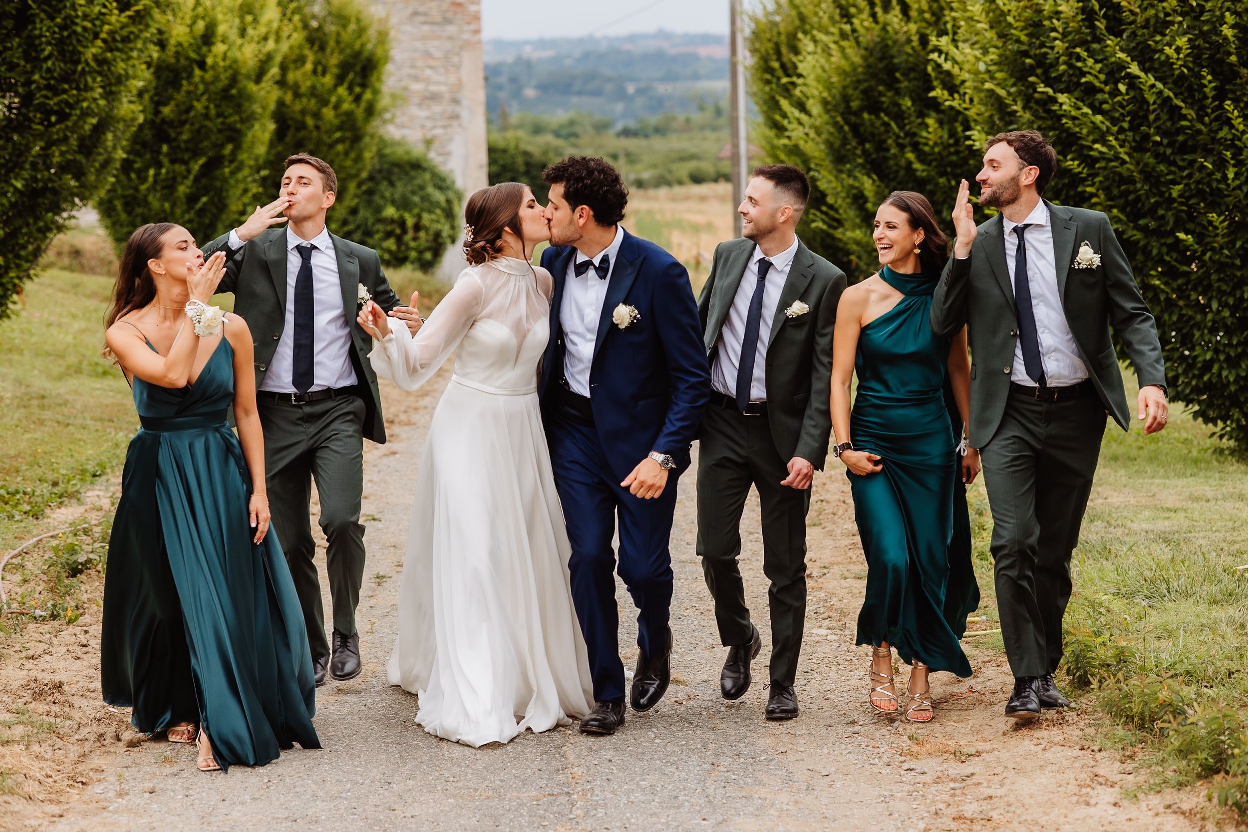 Wedding party in emerald green dresses and navy suits walking and laughing together on outdoor path with scenic view.