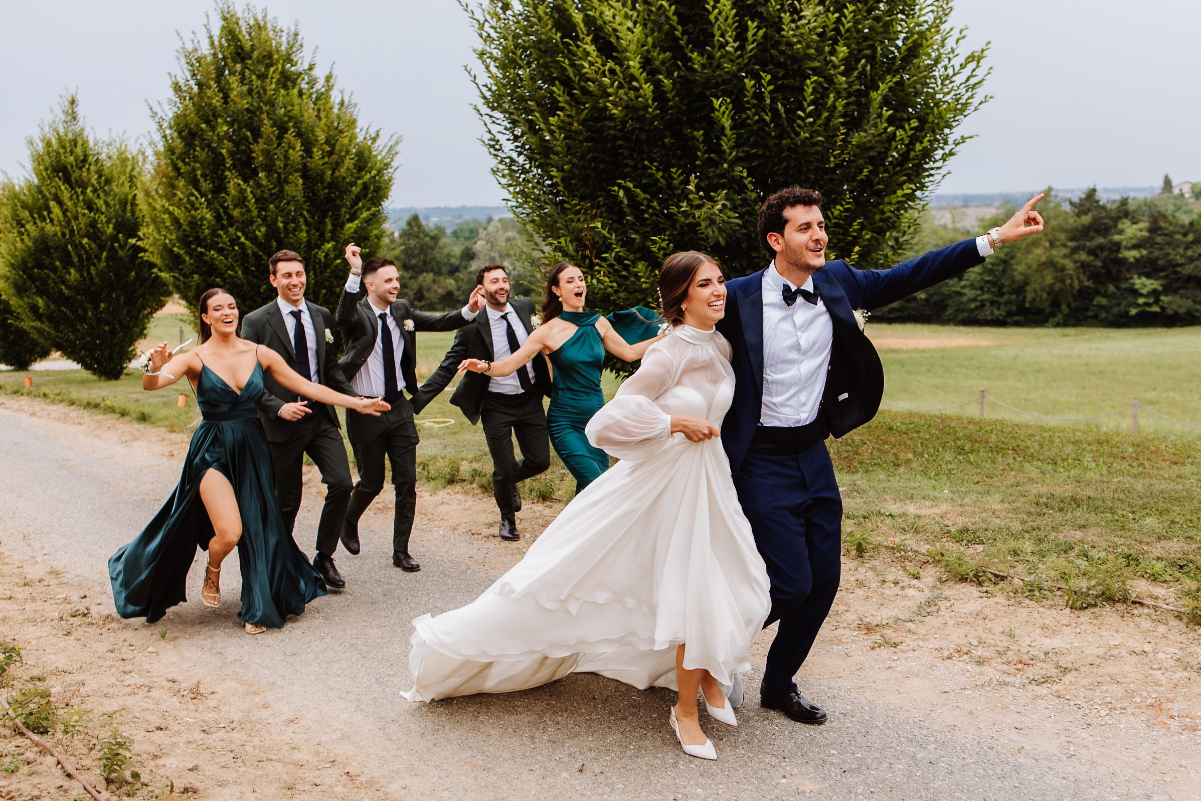 A wedding party in navy and green dresses walks together on a path while laughing and enjoying a sunny day outdoors.