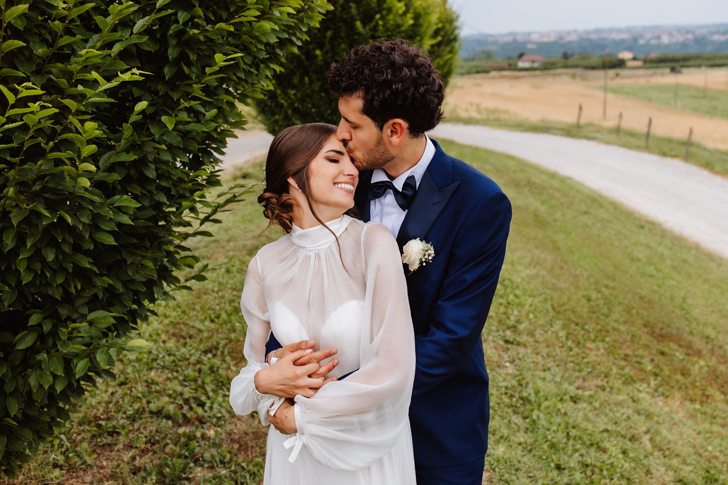 A romantic couple embraces on a countryside path next to green hedges on a sunny afternoon.