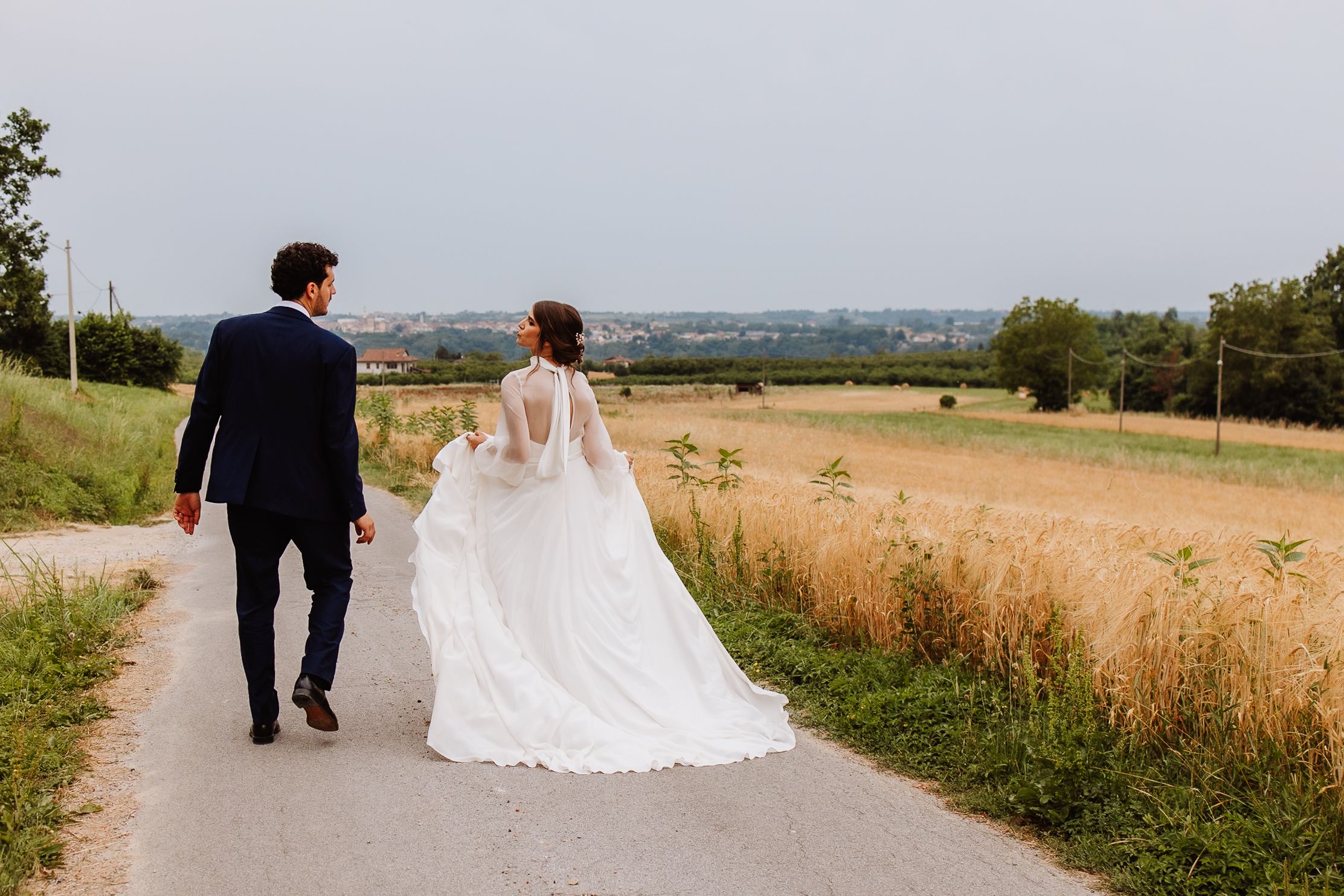 A newly married couple walks together along a scenic countryside path surrounded by golden summer fields.