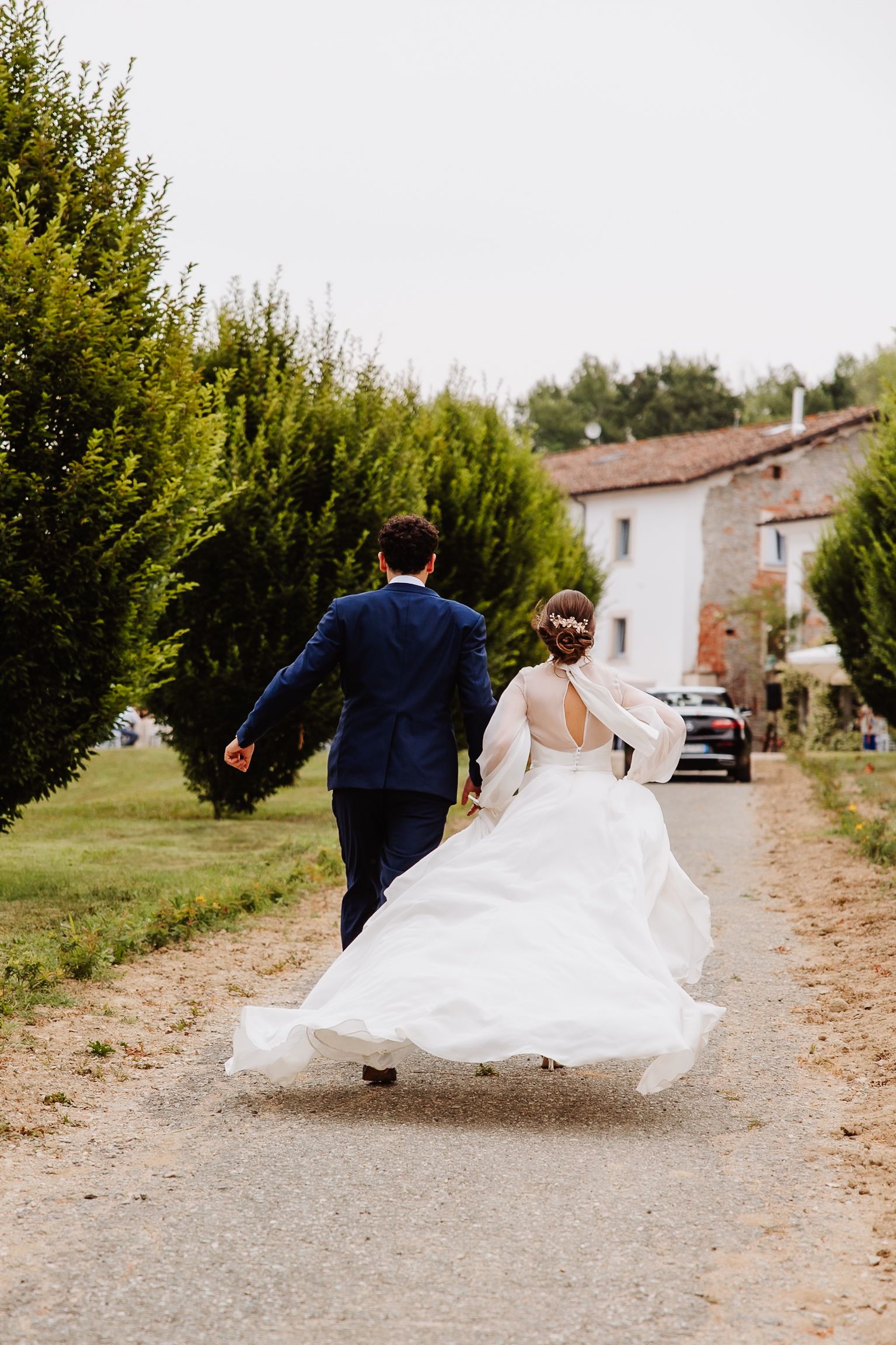 A couple in wedding attire walking down a rural path with the bride's white dress flowing in the breeze.