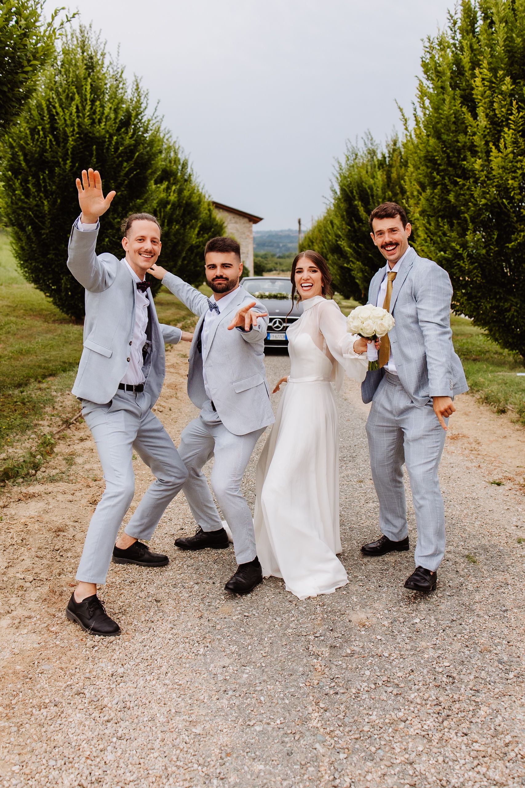 A wedding party in light grey suits strike fun poses together on a gravel path lined with evergreen trees.
