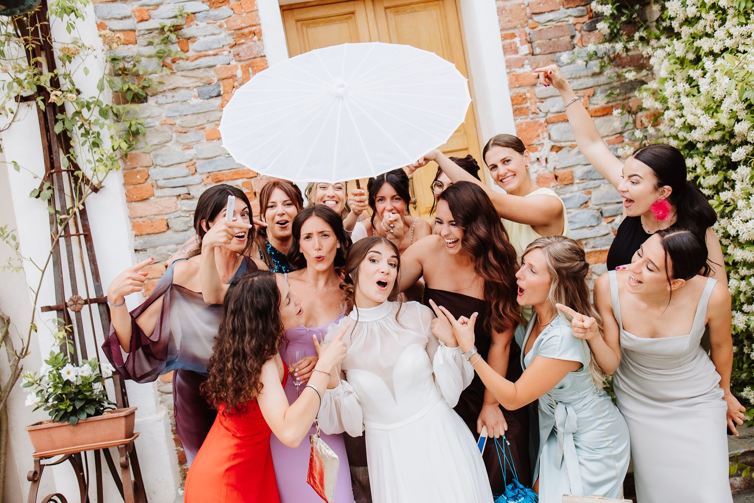 A group of elegantly dressed people share a white umbrella during a celebration on a sunny day by a brick wall.