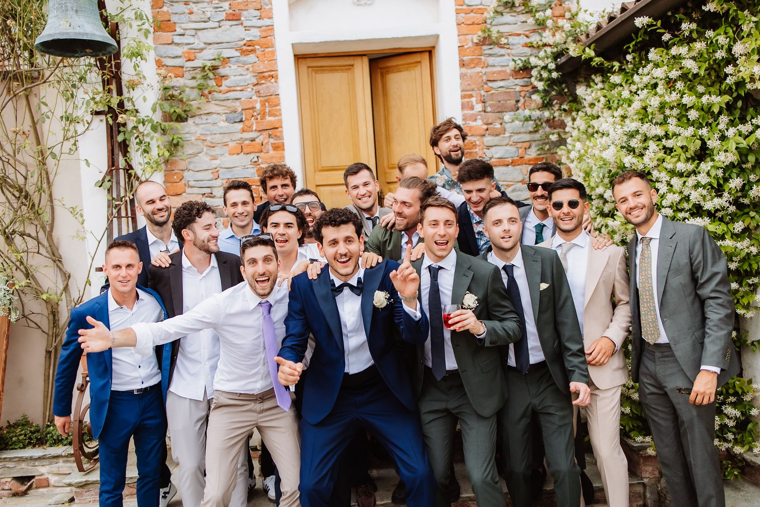 Groomsmen and wedding party enjoy drinks while standing in line against a rustic brick wall during outdoor reception.