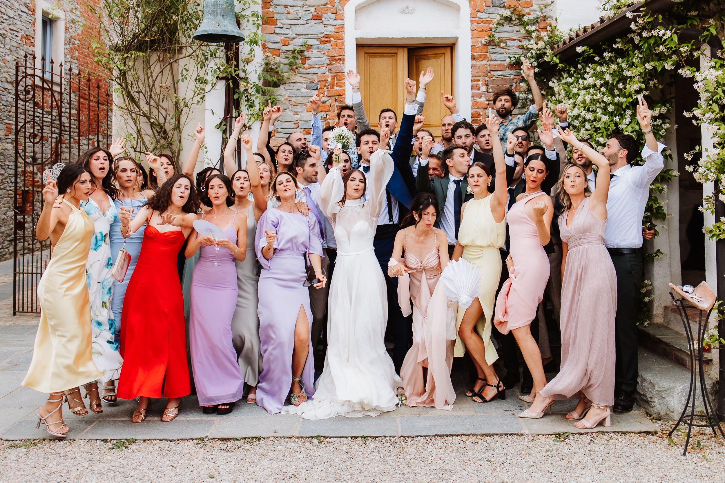A wedding party poses together outside a brick building in colorful pastel dresses and formal attire.