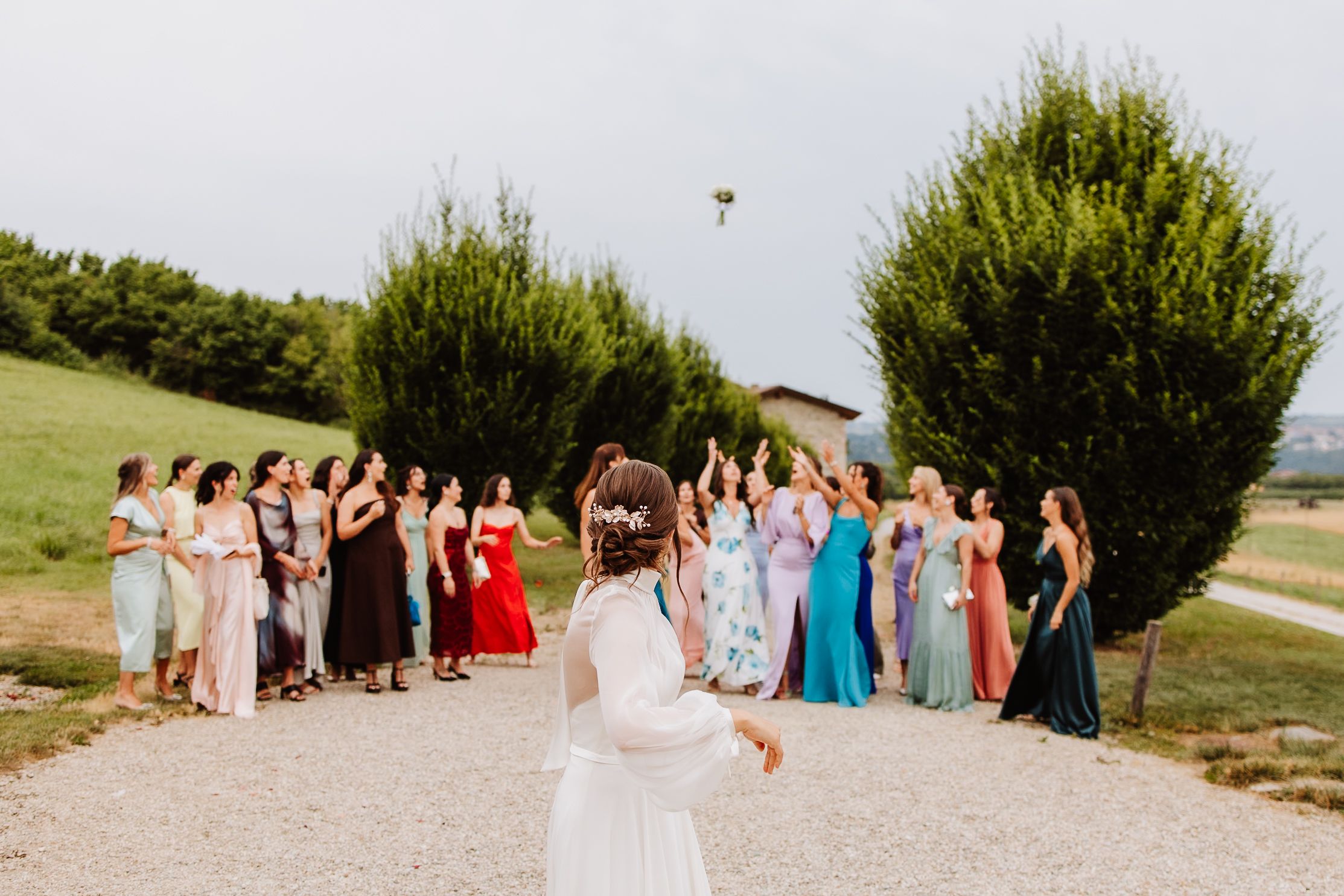 A wedding party gathers outdoors on a gravel path as someone tosses a bouquet into the air during a sunny celebration.