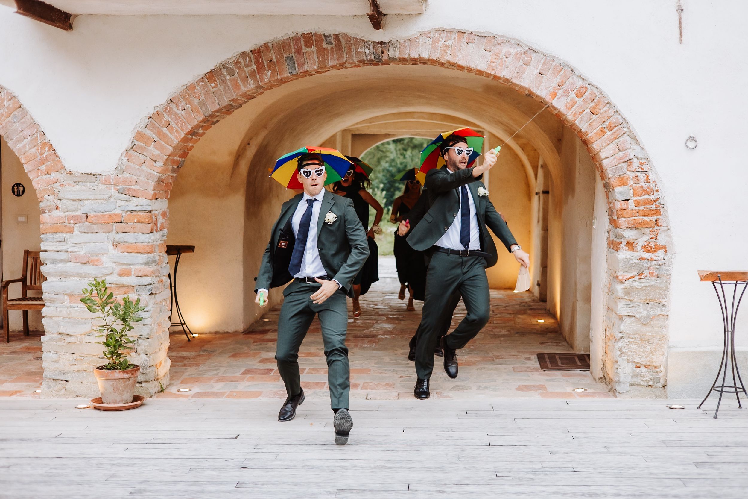 Two people in matching suits and blue masks jump playfully under an arched stone walkway.
