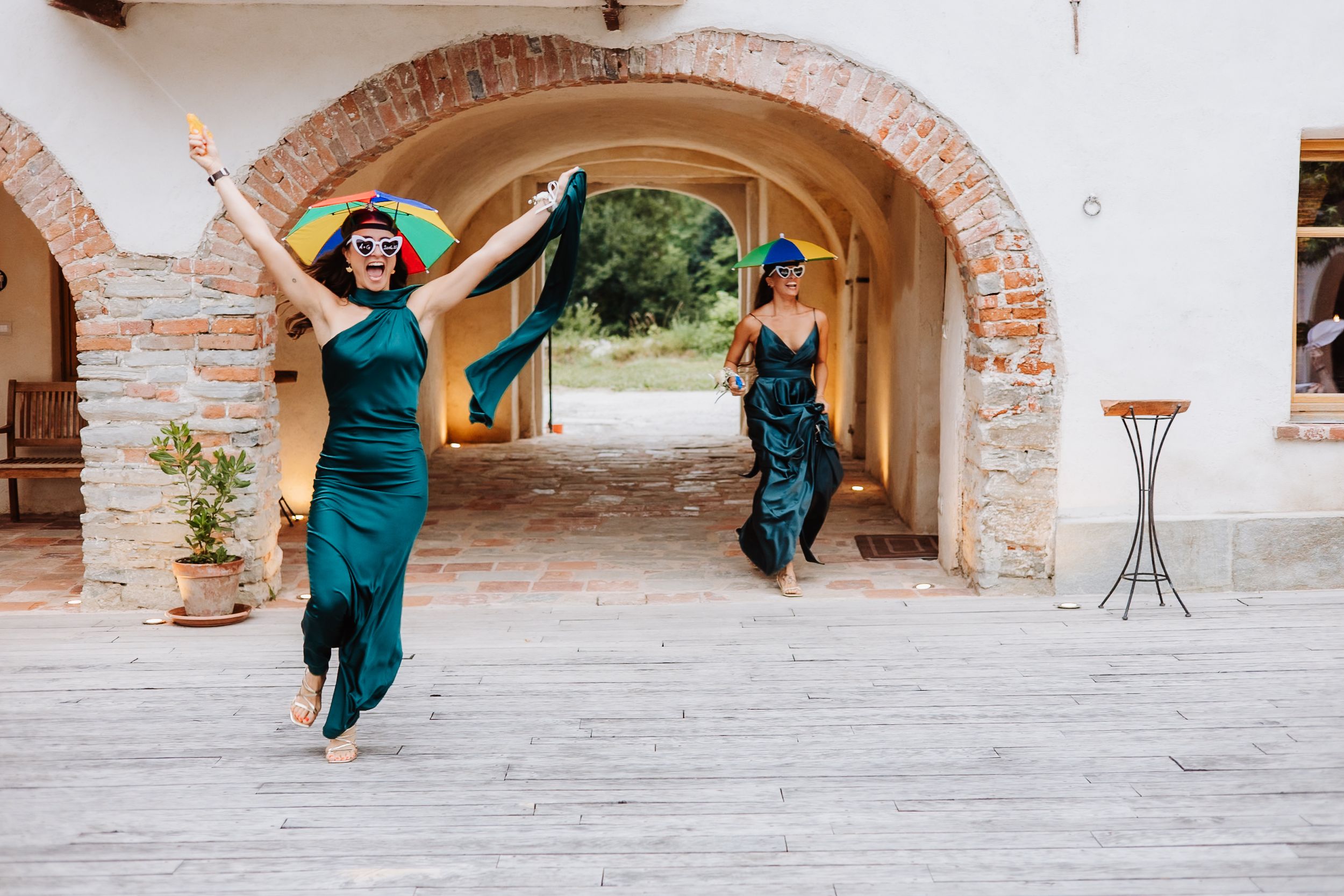 Two people in teal dresses jump joyfully under a stone archway in front of a rustic building.