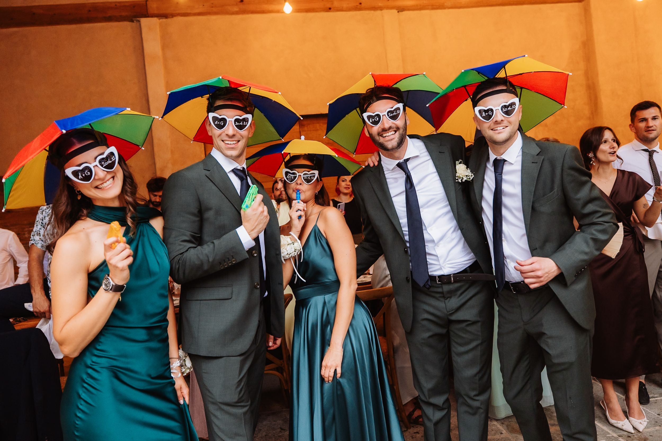 Wedding party guests celebrate with colorful rainbow umbrellas while wearing formal suits and dresses at reception.